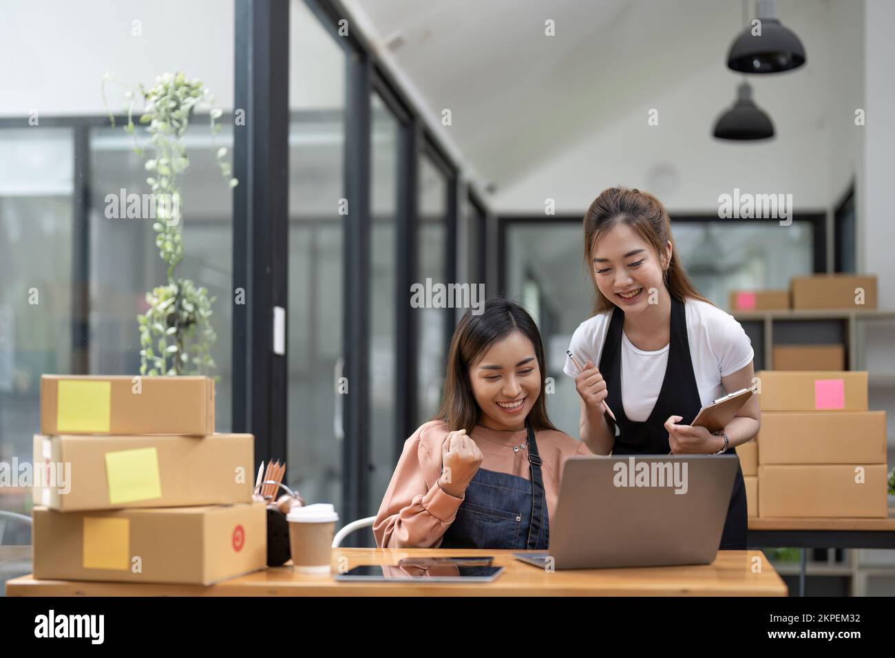 Fröhliche zwei asiatische Unternehmerinnen, lächeln für Verkaufserfolg, nachdem Sie die Bestellung im Online-Shop mit einem Smartphone im Heimbüro geprüft haben, Konzept von Stockfoto