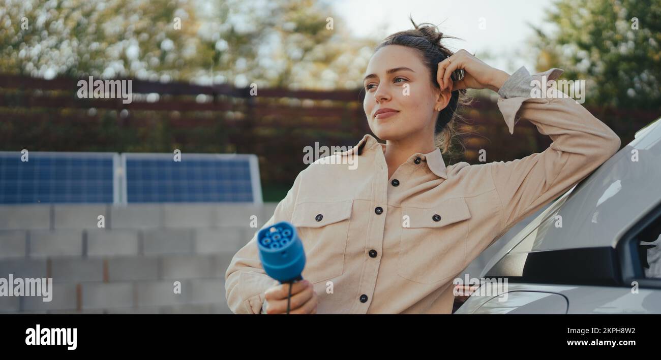 Junge Frau, die das Stromkabel von ihrem Elektroauto hält, bereit, es zu Hause zu laden, ein nachhaltiges und wirtschaftliches Transportkonzept. Stockfoto