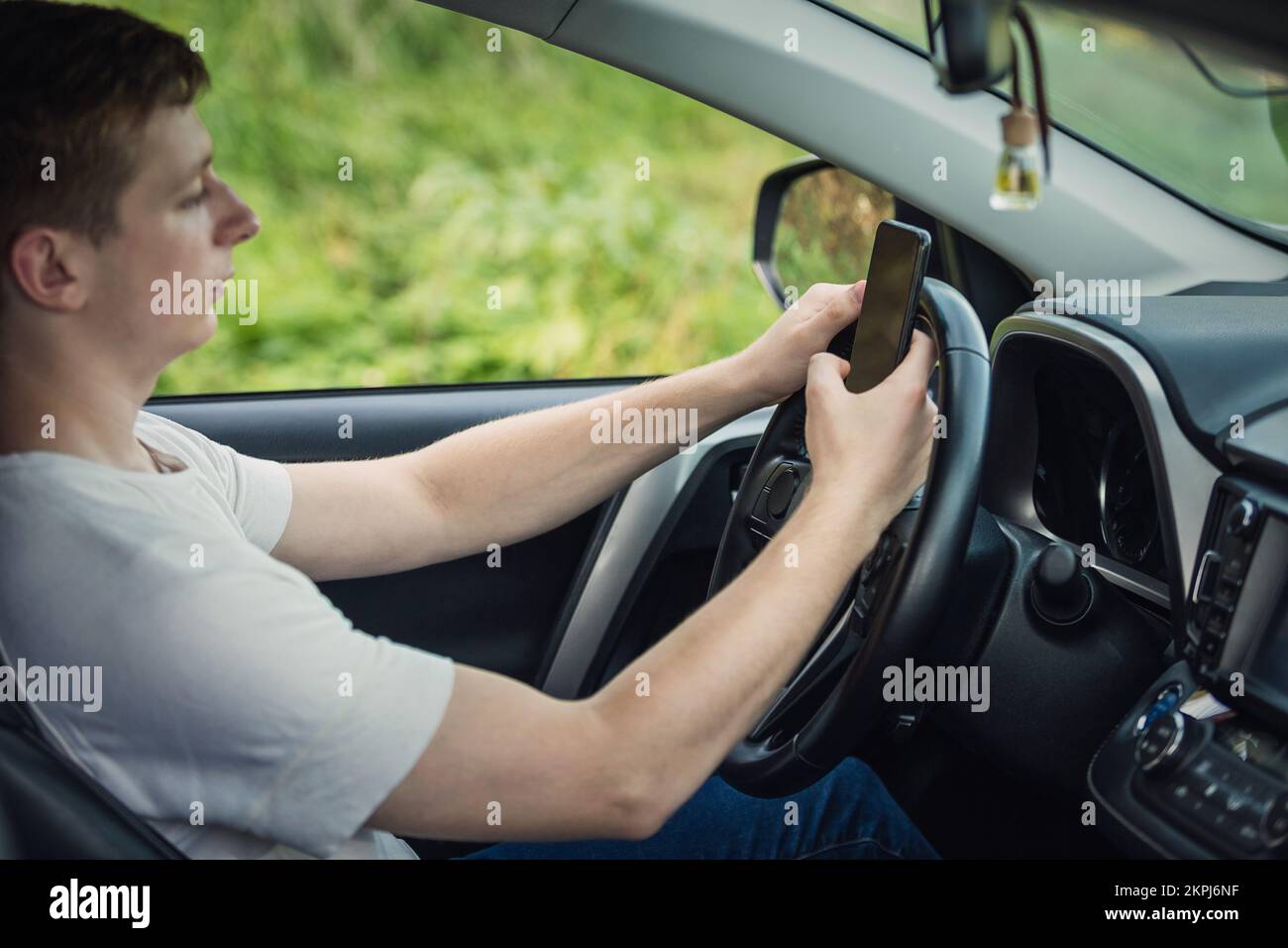Ein junger Fahrer, der sich vor dem Lenkrad von seinem Telefon abgelenkt hat und sein Smartphone während der Fahrt mit einer Hand benutzte. Risiko- und Gefahrensituation Stockfoto