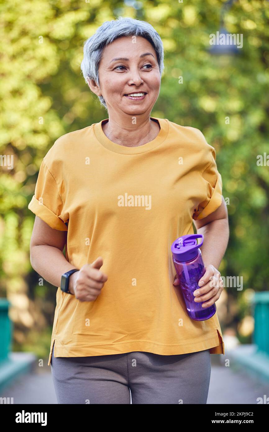 Eine reife, sportliche Asiatin, die eine Flasche Wasser in der Hand hält, trainiert im Freien und joggt im Sommerpark. Gesunde aktive Lebensweise der Rentner, Stockfoto