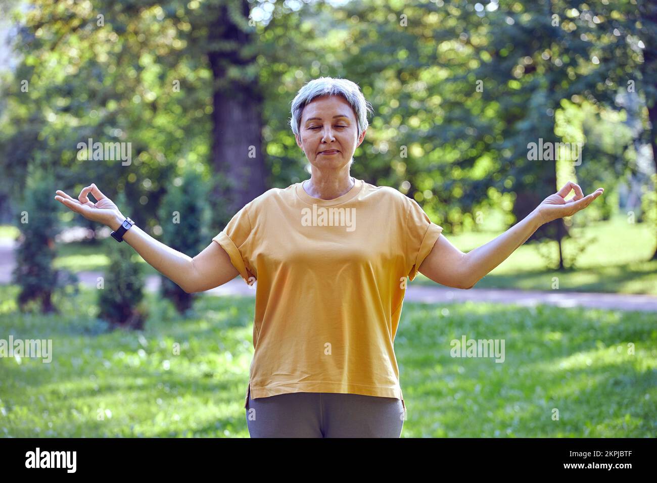Eine ältere, einsame Frau mit geschlossenen Augen sorgt dafür, dass Mudra-Geste den neuen Tag beginnt und sich beim Training lebendig anfühlt, lächelt allein im Sommerpark auf grünen Bäumen im Hintergrund Stockfoto