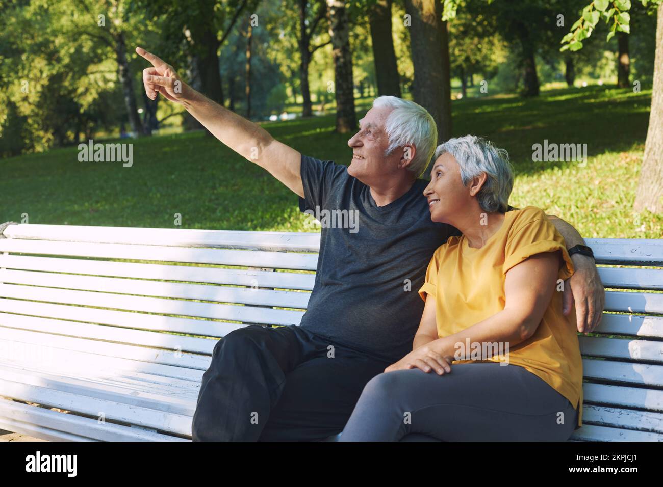 Positives multi-rassisches älteres 60s-Pärchen ruht sich nach einem Spaziergang auf einer Bank im Park aus, der Ehemann zeigt den Zeigefinger nach oben und spricht mit einer lächelnden Frau, die sich entspannt Stockfoto