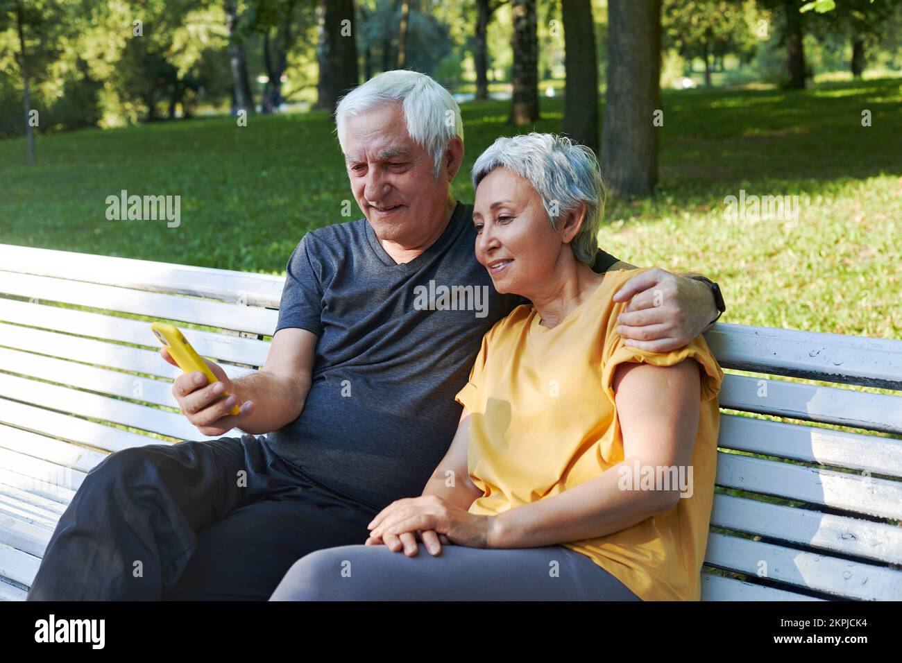 Ältere Ehegatten sitzen im Sommerpark auf einer Bank mit Smartphone. Grauhaarige Paare schauen auf Handybildschirm Genießen Sie neue Anwendung Outdoor nutzen moderne Technologie Stockfoto