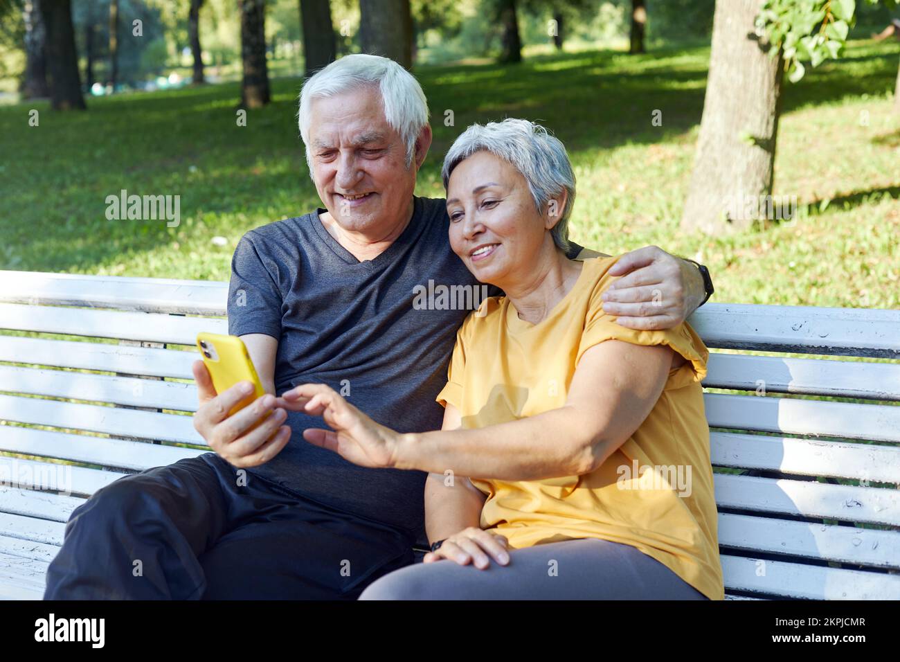 Ältere Ehepartner sitzen auf einer Bank im Sommerpark mit Smartphone. Grauhaarige Paare schauen auf Handybildschirm Genießen Sie neue Anwendung Outdoor nutzen moderne Technologie Stockfoto