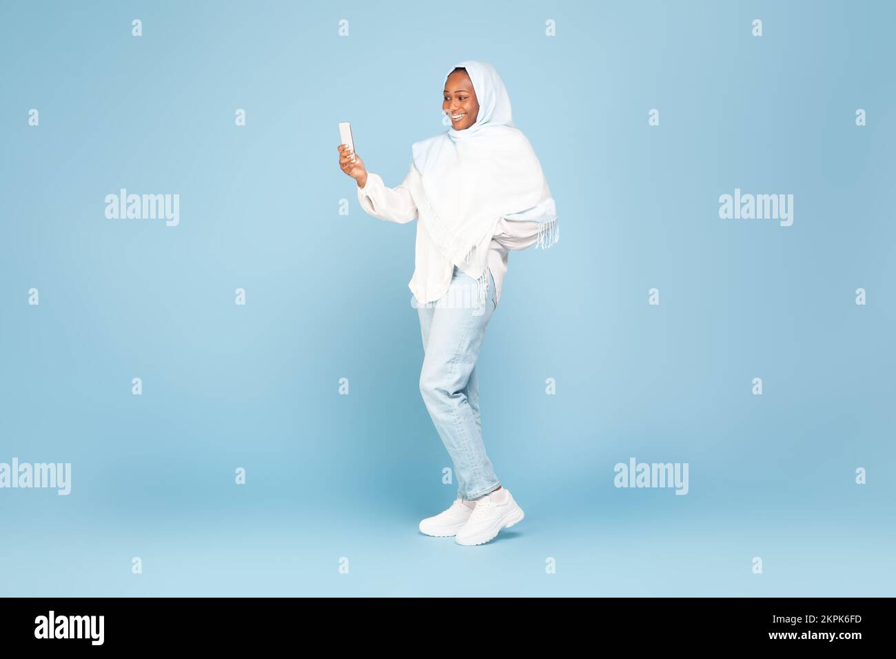 Glückliche afroamerikanische Frau in Hidschab, die Handy benutzt, auf blauem Studiohintergrund steht, freier Platz, volle Körperlänge Stockfoto