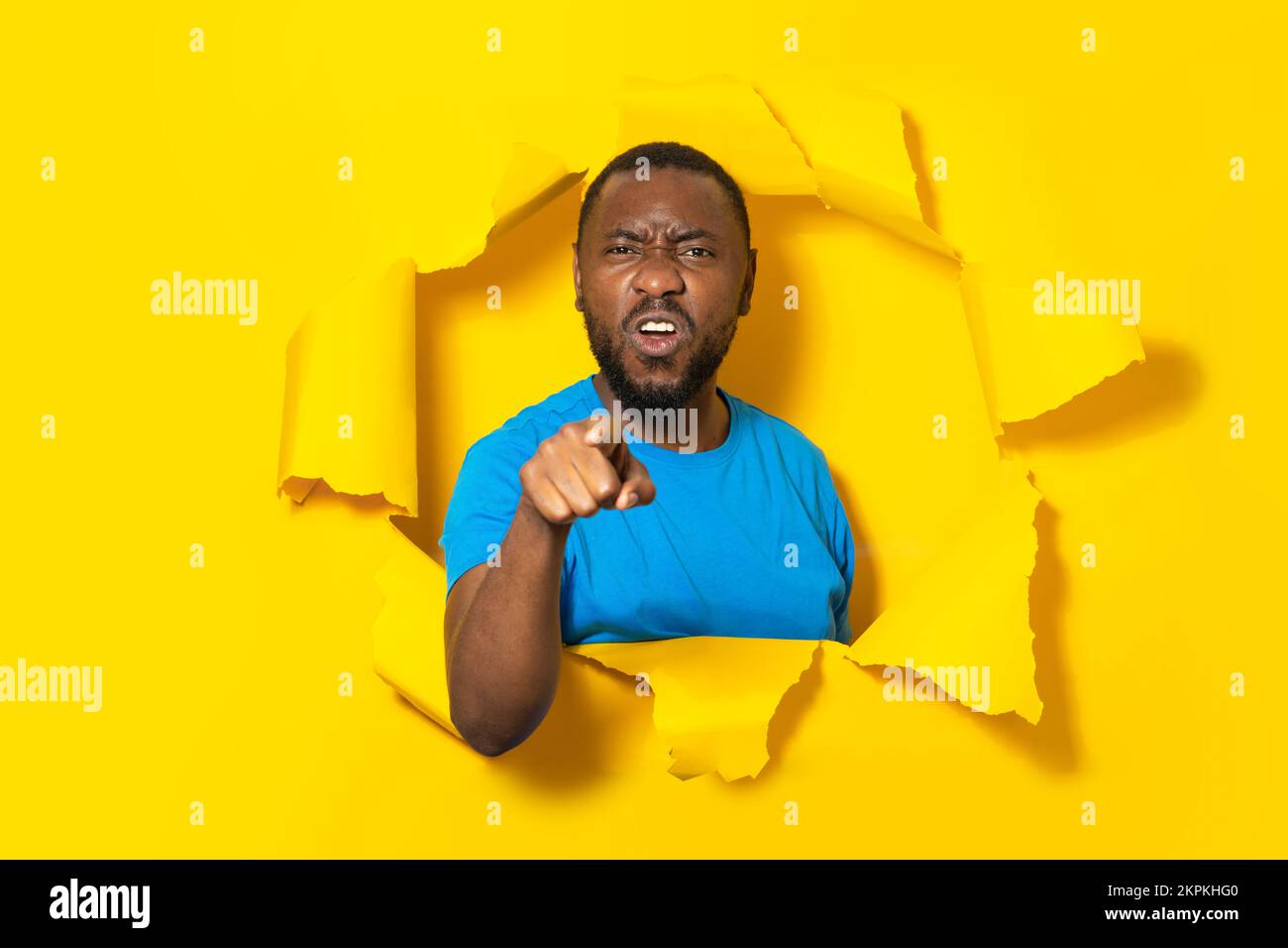 Ein aggressiver Schwarzer, der mit dem Finger auf die Kamera zeigt, dich beschuldigt, in einem Loch in zerrissenem gelben Papier posiert Stockfoto