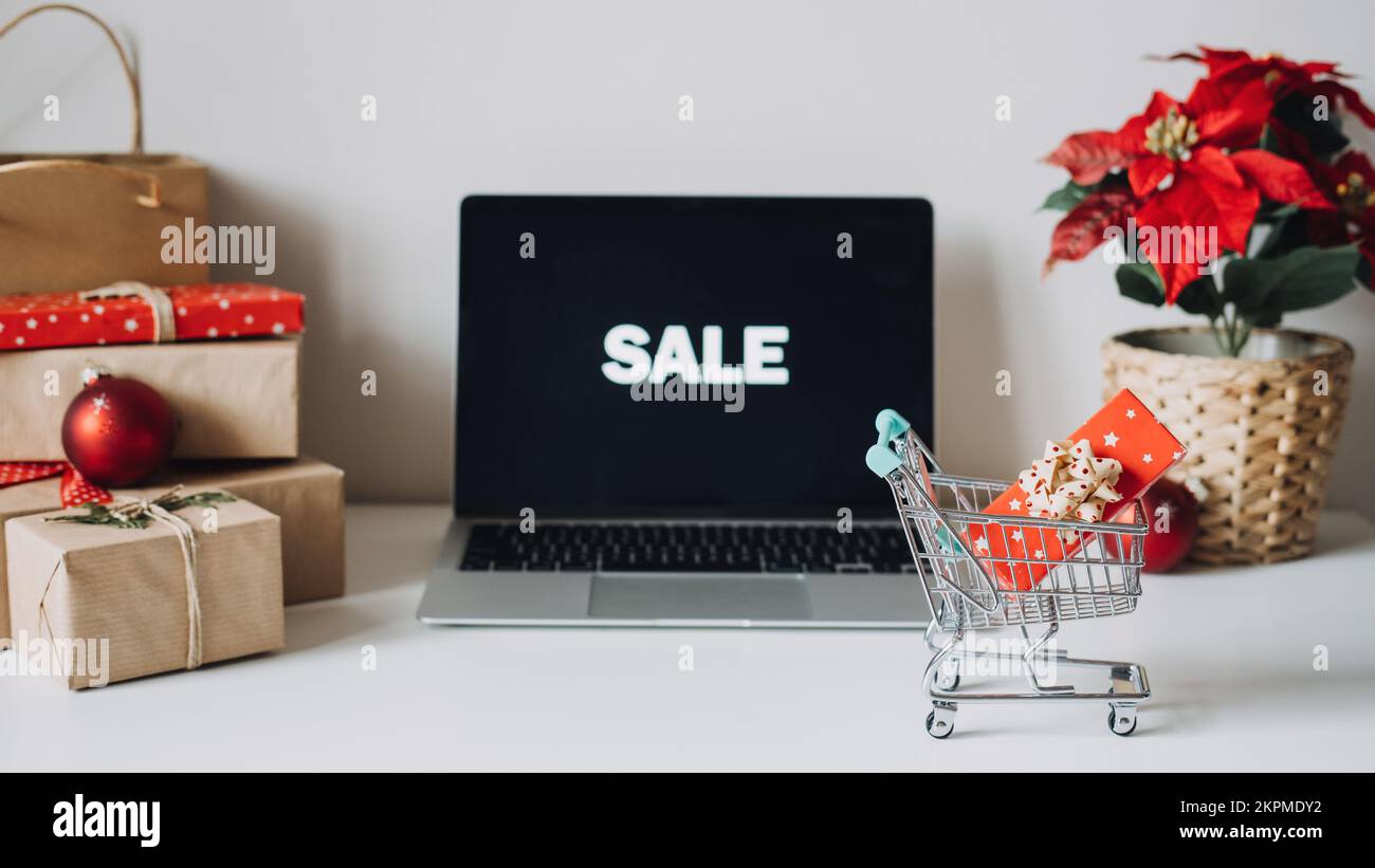 Erfolgreiche Weihnachtszeit im E-Commerce, weihnachtliches Online-Shopping. Offener Laptop-Bildschirm, Mini-Einkaufswagen, Poinsettias Christmas Stockfoto