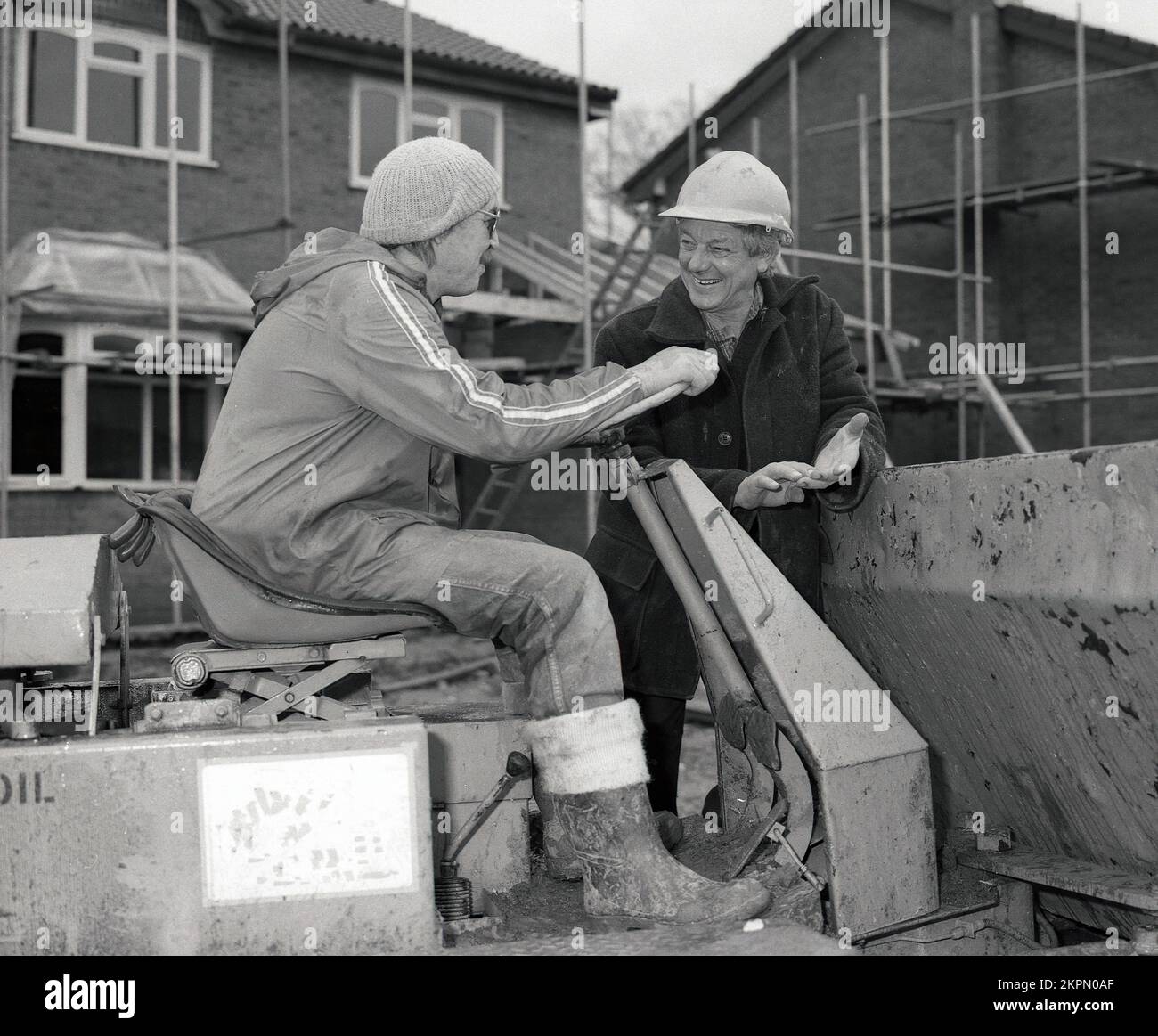 1980er, draußen auf einer Baustelle, zwei Arbeiter, die ein Gespräch führen, einer, möglicherweise ein Vorarbeiter mit Schutzhelm, und ein Arbeiter mit Bommelhelm, der auf einem Kippkipper sitzt, einem Baufahrzeug, das eine Art Kipplaster ist, England, Großbritannien. Stockfoto