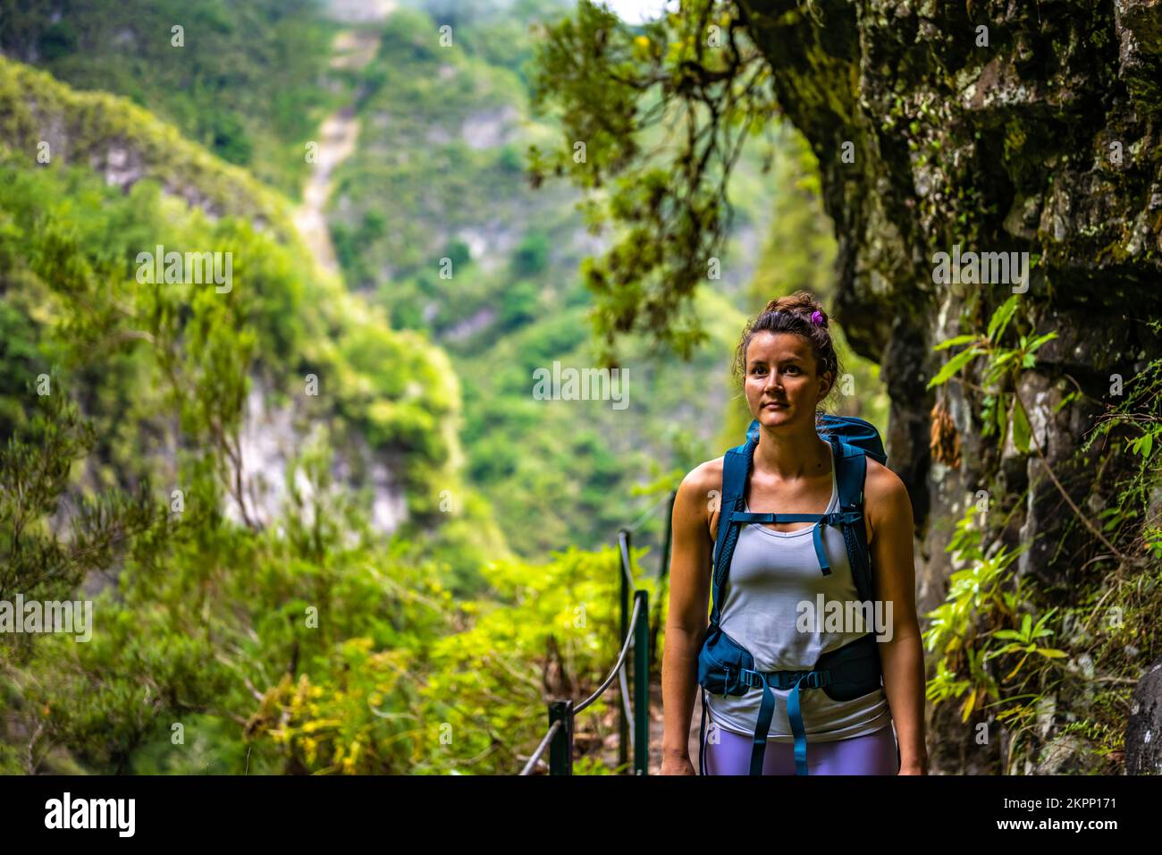 Beschreibung: Eine sportliche Frau mit Rucksackblick in die Ferne vom abenteuerlichen Dschungelweg entlang eines grünen, überwucherten Kanals. Levada von Caldeir Stockfoto