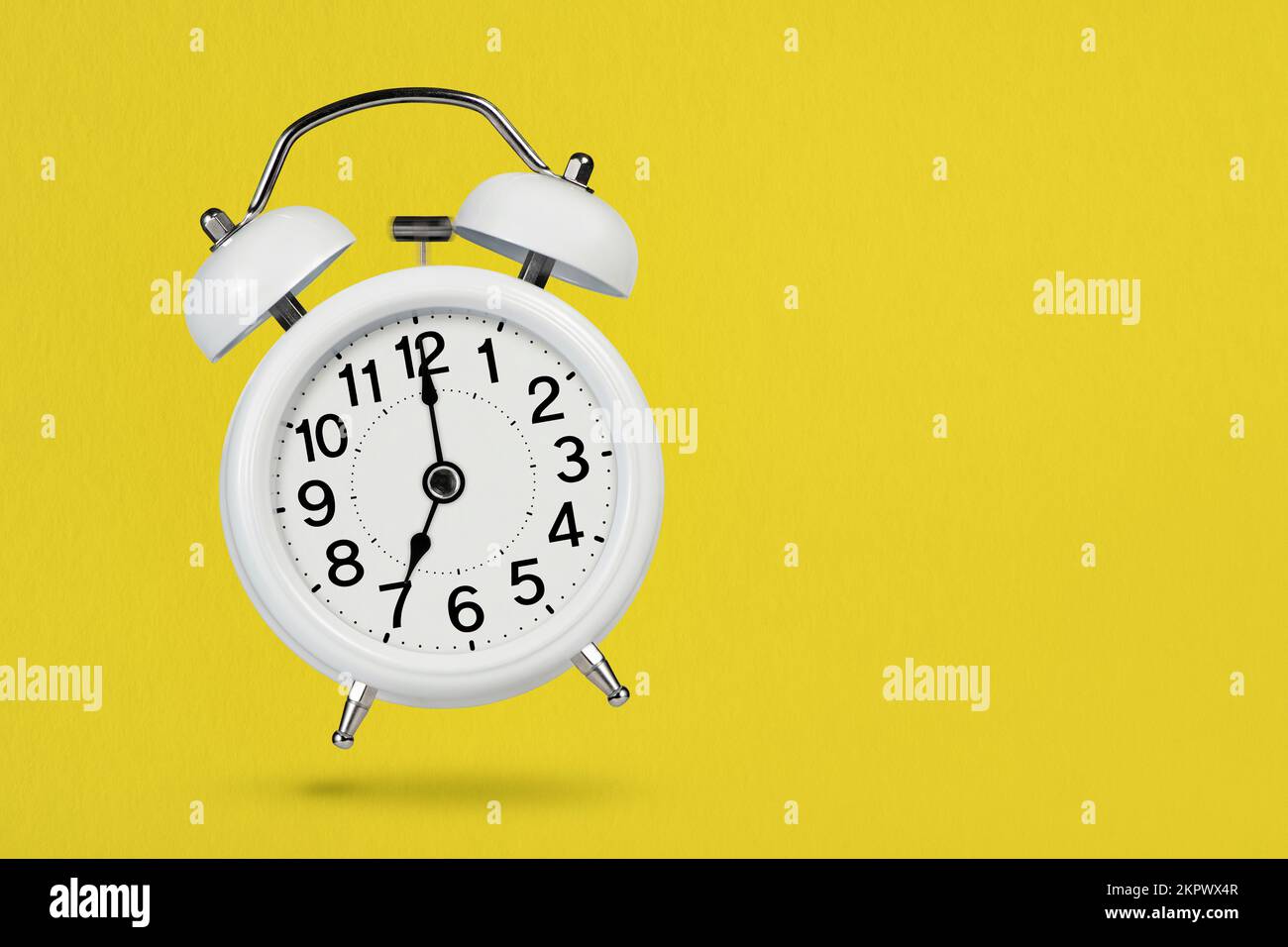 Ein weißer Vintage-Wecker zeigt 7 Uhr an und wirft einen Schatten auf einen gelben Hintergrund. Das Konzept des Morgen- und Frühaufwachens. Zeit zu spielen oder zu spielen Stockfoto
