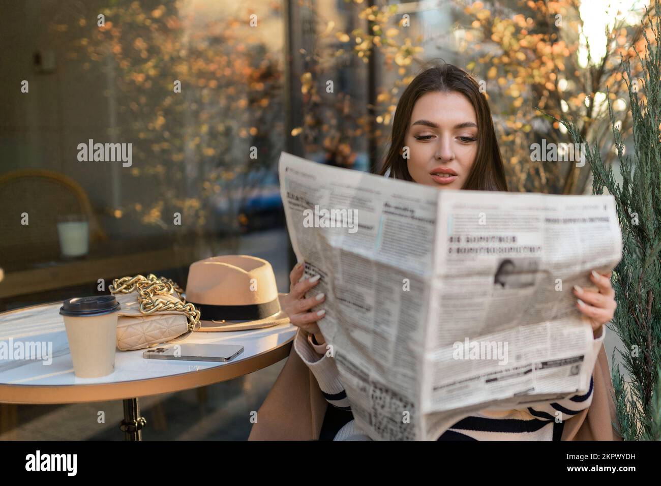 Ein hübsches Mädchen im Mantel liest eine Zeitung und lächelt, während es sich in einem Café im Freien entspannt Stockfoto