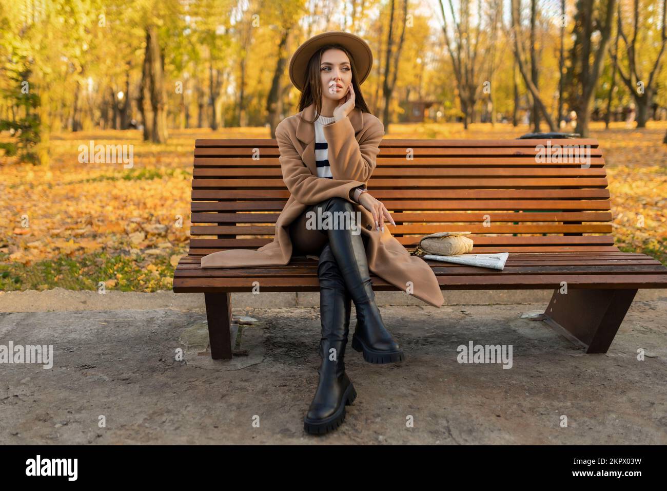 Porträt eines attraktiven, stilvollen, lächelnden Porträts einer attraktiven, stilvollen, lächelnden Frau mit langen Haaren, die im Park spaziert, gekleidet in einem warmen braunen Mantel Stockfoto