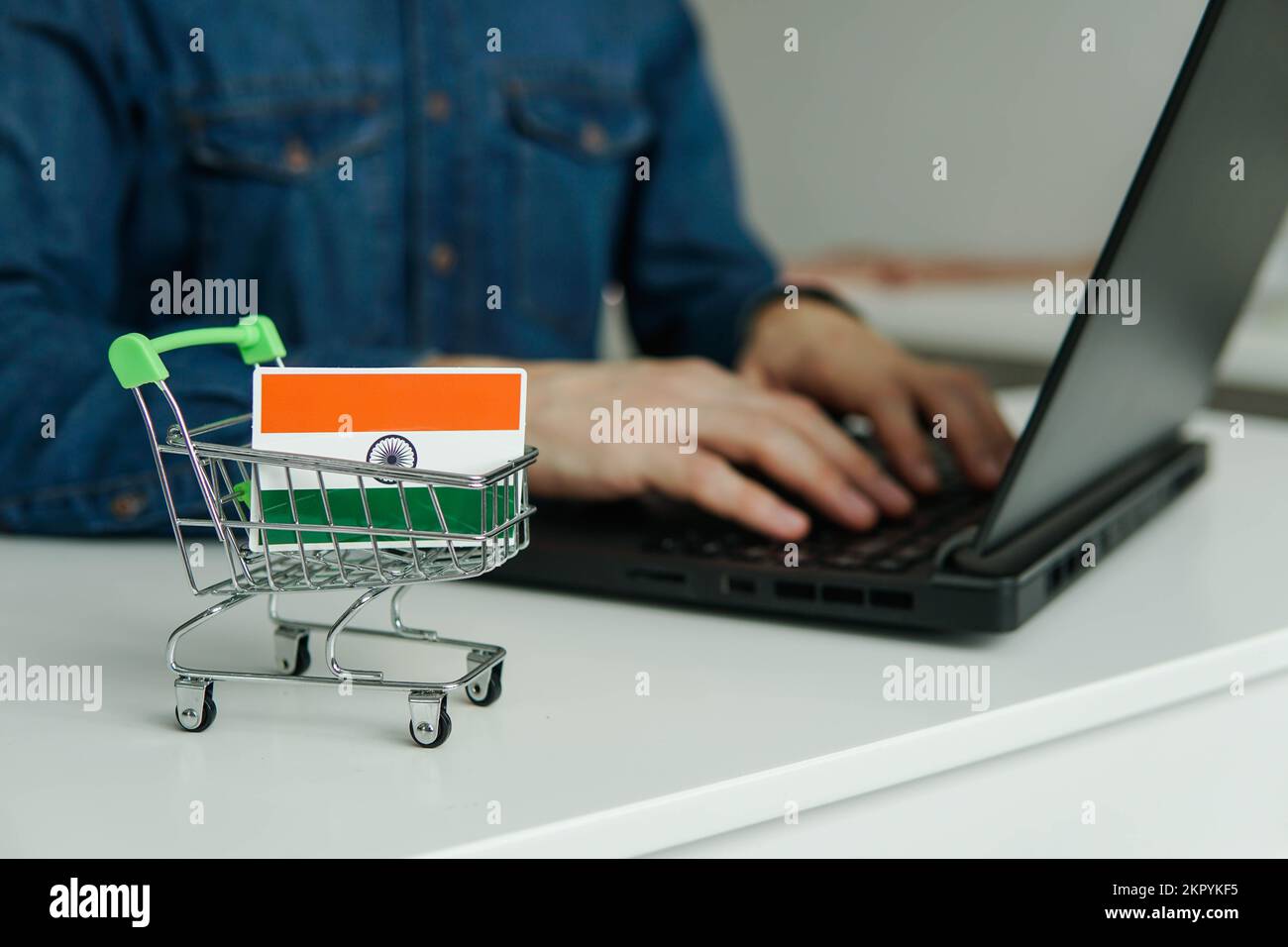 Kleiner Einkaufswagen mit indischer Flagge auf dem Tisch. Mann, der ein Notebook zum Einkaufen im Internet verwendet. Stockfoto