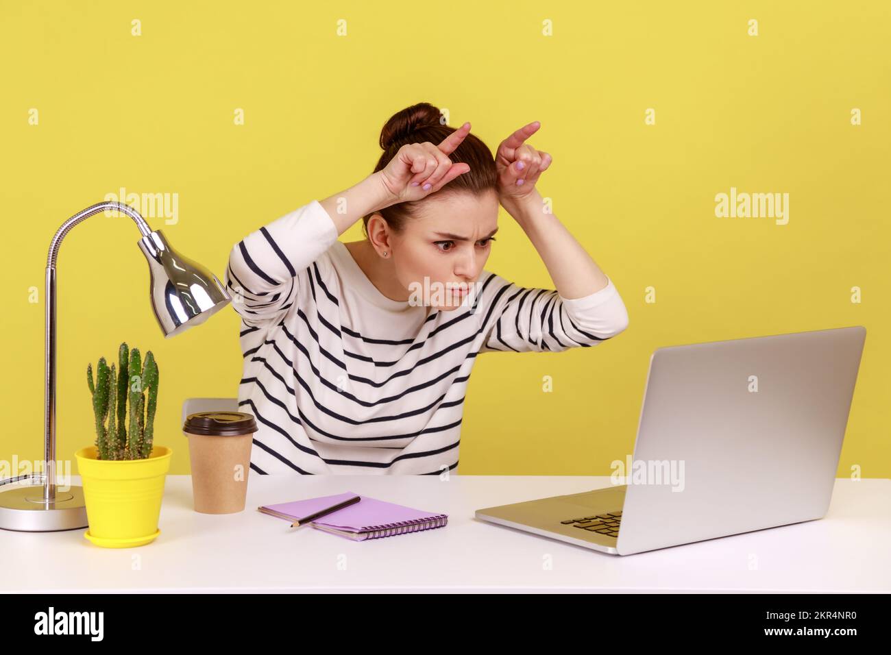 Eine wütende, herrisch besetzte Sekretärin, die Finger über dem Kopf hält und Bullenhörner am Arbeitsplatz zeigt, die drohen, wütend auf den Laptop-Bildschirm zu schauen. Studioaufnahmen im Innenbereich isoliert auf gelbem Hintergrund. Stockfoto