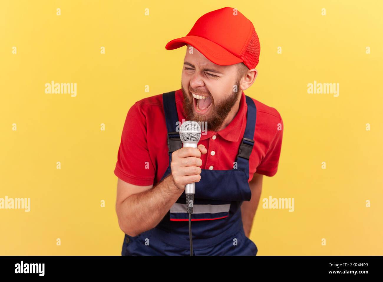 Portrait eines extrem aufgeregten Arbeiters in blauer Uniform und roter Mütze, der sich auf einer Firmenfeier entspannt und Karaoke auf der Arbeit singt. Innenstudio-Aufnahme isoliert auf gelbem Hintergrund. Stockfoto