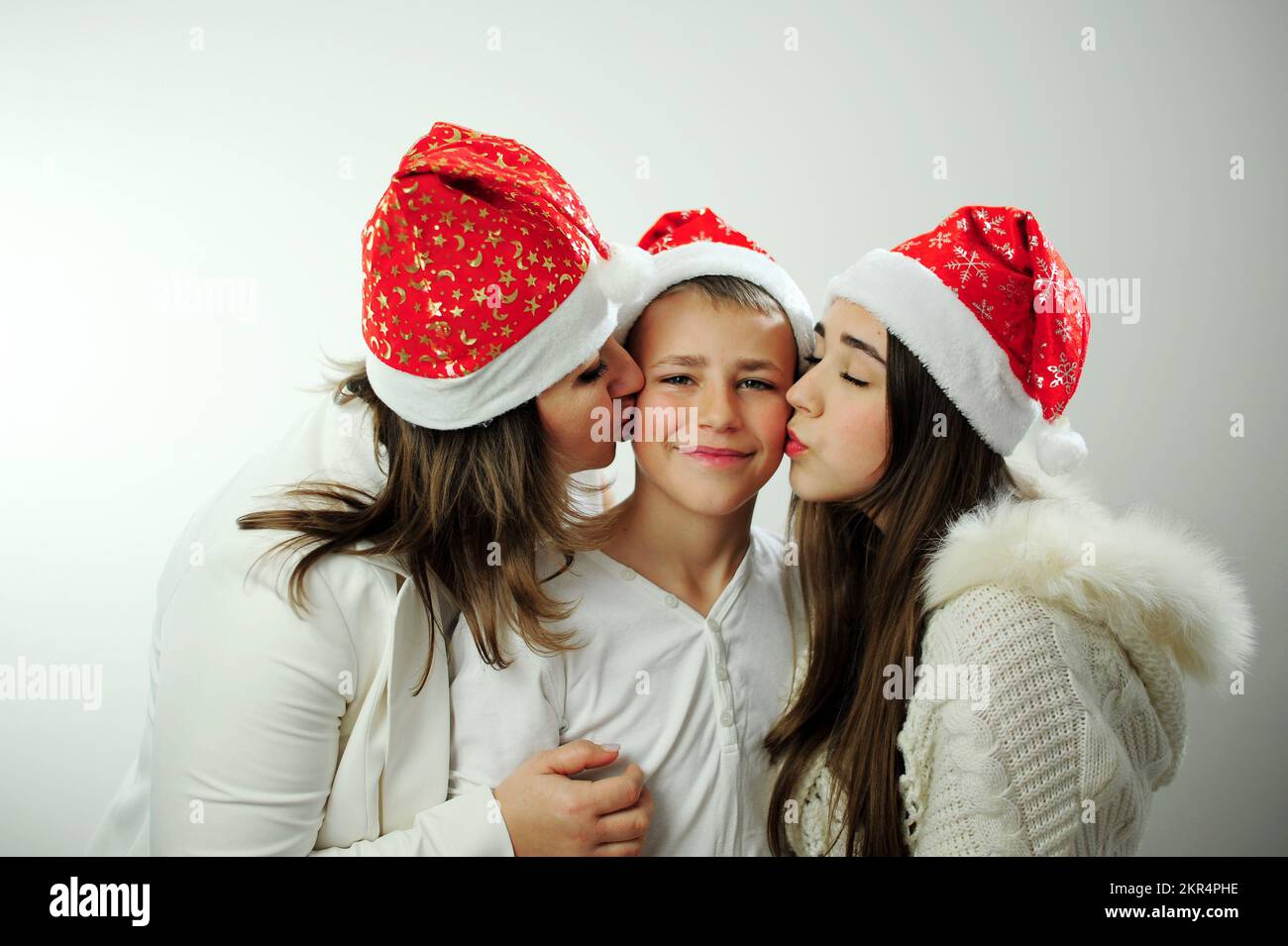 Mutter, Tochter und Sohn feiern Neujahr 3 Menschen sind in Weiß gekleidet, tragen Silvester-rote Hüte, unvollständige Familie umarmt mutter und Tochter zwei Seiten ihres Sohnes und Bruders Stockfoto