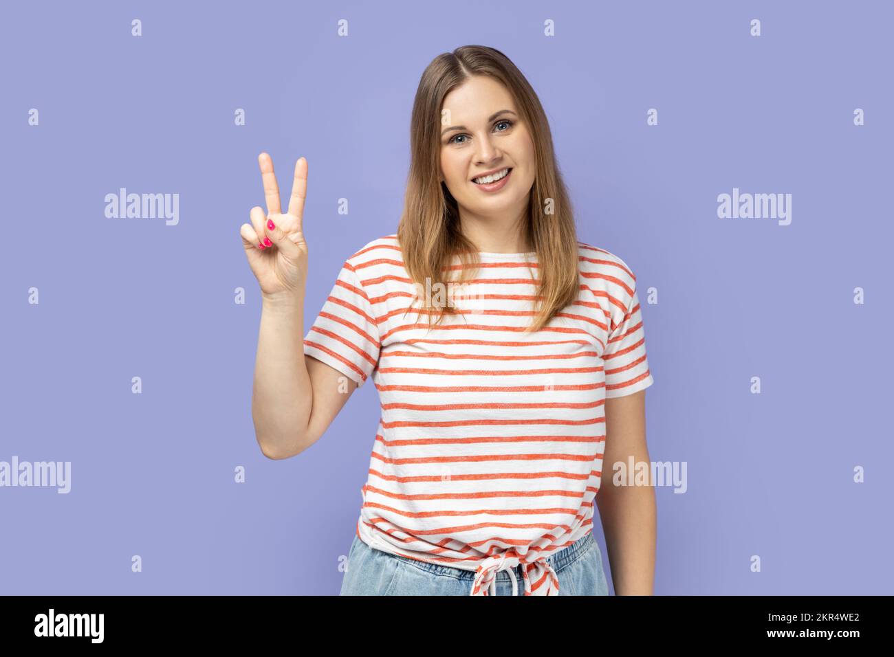 Porträt einer schönen blonden Frau, die ein gestreiftes T-Shirt trägt, fröhlich lächelt und ein V-Zeichen oder eine Friedensgeste zeigt, um sich über den Sieg zu freuen. Studio-Aufnahme im Innenbereich isoliert auf lila Hintergrund. Stockfoto