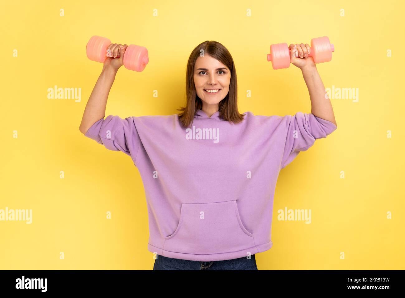 Sportliche Frau erhob die Arme, machte Fitnessübungen mit pinkfarbenen Kurzhanteln, trainierte Hände, schaute mit einem Lächeln in die Kamera, trug einen lila Hoodie. Innenstudio-Aufnahme isoliert auf gelbem Hintergrund. Stockfoto