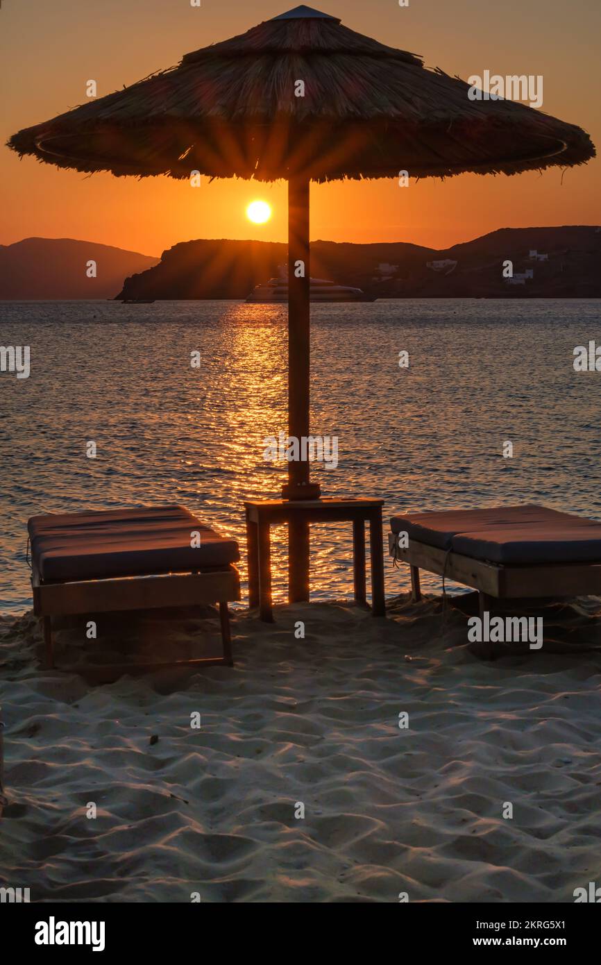 Blick auf eine Sonnenliege, einen Sonnenschirm und einen atemberaubenden orangefarbenen Sonnenuntergang am Strand von Mylopotas in iOS Griechenland Stockfoto