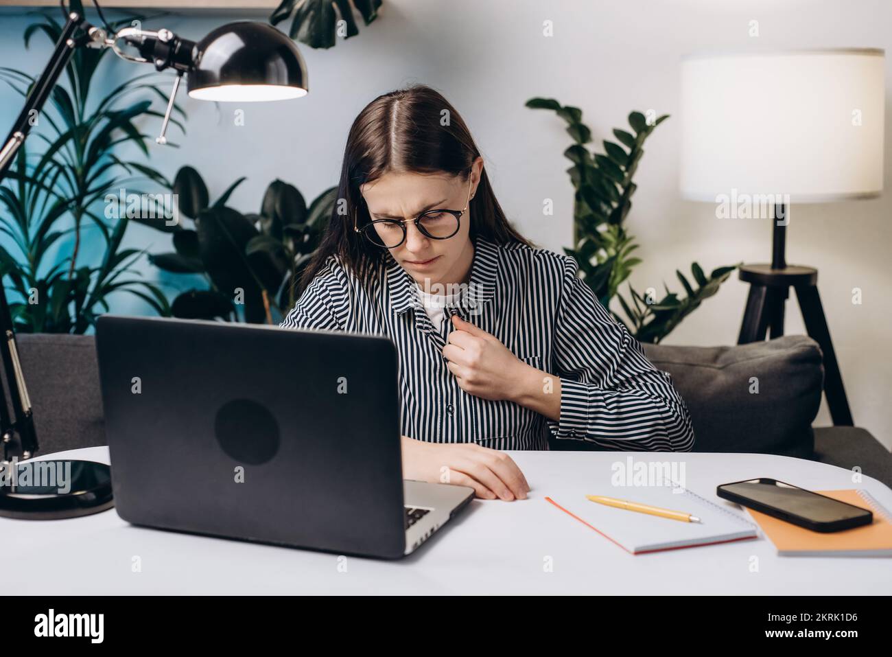 Gestresste junge Frau mit Brille, die einen Herzinfarkt hat, sitzt auf der Couch im Heimbüro. Arbeitskraft erschöpft vom Computer am Arbeitsplatz mit Stress Stockfoto