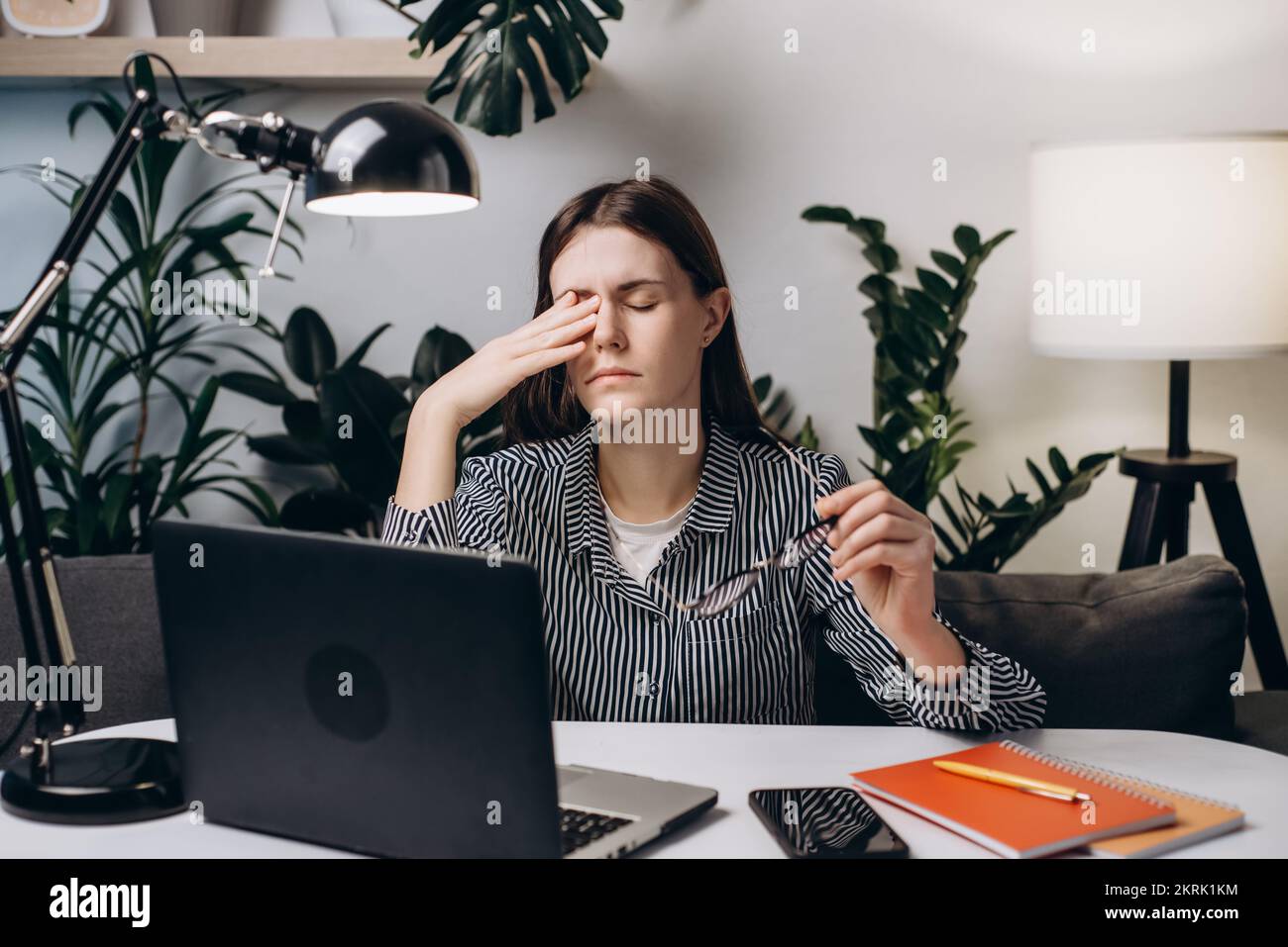Junge Frau, die im Heimbüro auf dem Sofa sitzt, arbeitet auf einem Laptop und hält die Brille fest. Massage Nasensteg reduziert die Belastung der Augen. Trockene Augen, Laser-Sehkorrektur Stockfoto