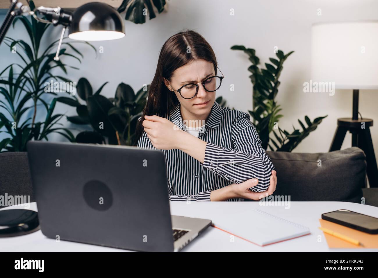 Nahaufnahme einer traurigen jungen Frau mit Brille, die am Arbeitsplatz auf dem Sofa sitzt und Schmerzen und Beschwerden im Ellenbogengelenk erlebt Stockfoto