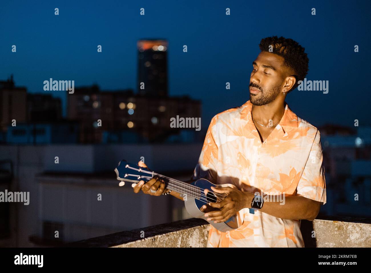 Positiver afroamerikanischer Gitarrist, der Musik auf Ukulele spielt, während er ein Lied probt, das gegen die Nachtstadt steht Stockfoto