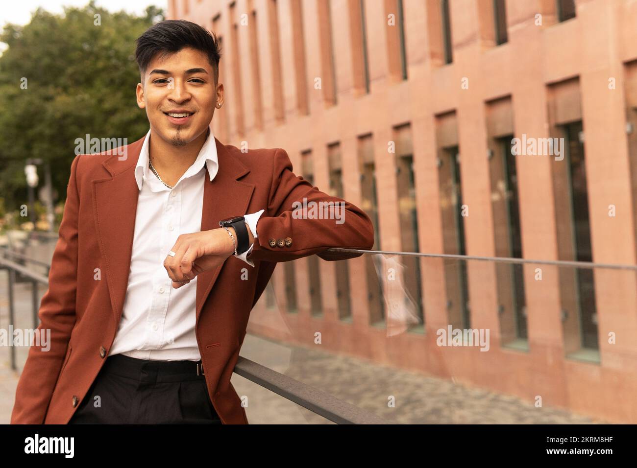 Positiver, ethnischer Unternehmer, der einen eleganten Anzug trägt, sich auf einen Glaszaun lehnt, in die Kamera schaut und lächelt Stockfoto