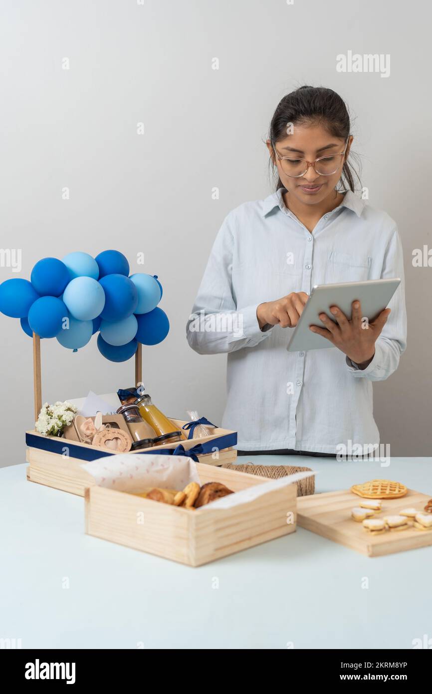 Positive junge Frau in legerem Outfit, die sich ein Tablet angesehen hat, während sie neben dem Tisch stand, mit Geschenkboxen mit dekorierten blauen Ballons Stockfoto