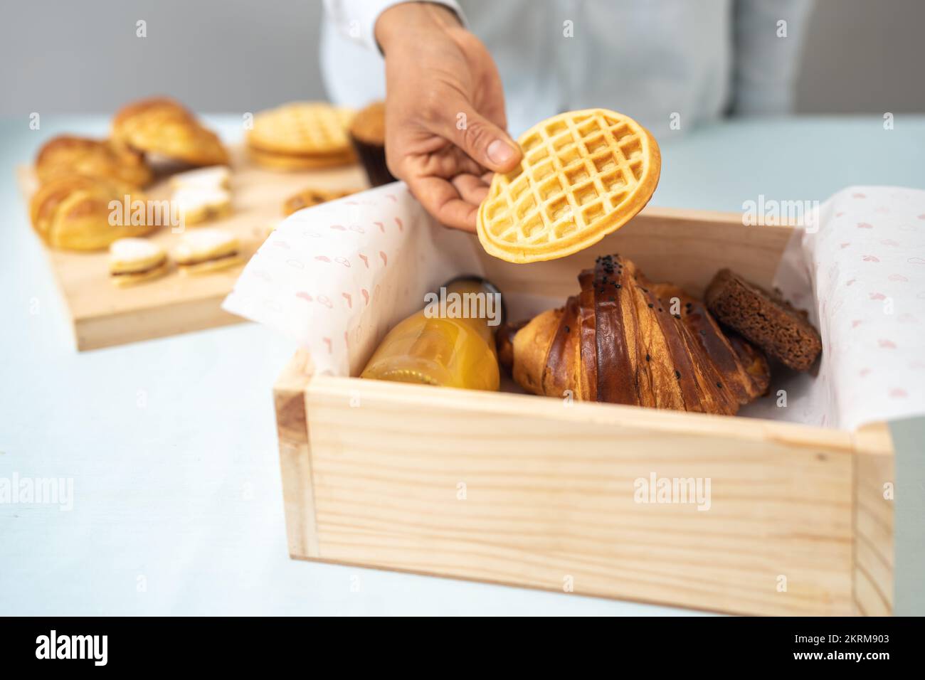 Eine anonyme Frau, die köstliche, hausgemachte Waffeln in eine Holzbox mit frischen Getränkekekekeksen und Croissants packt Stockfoto