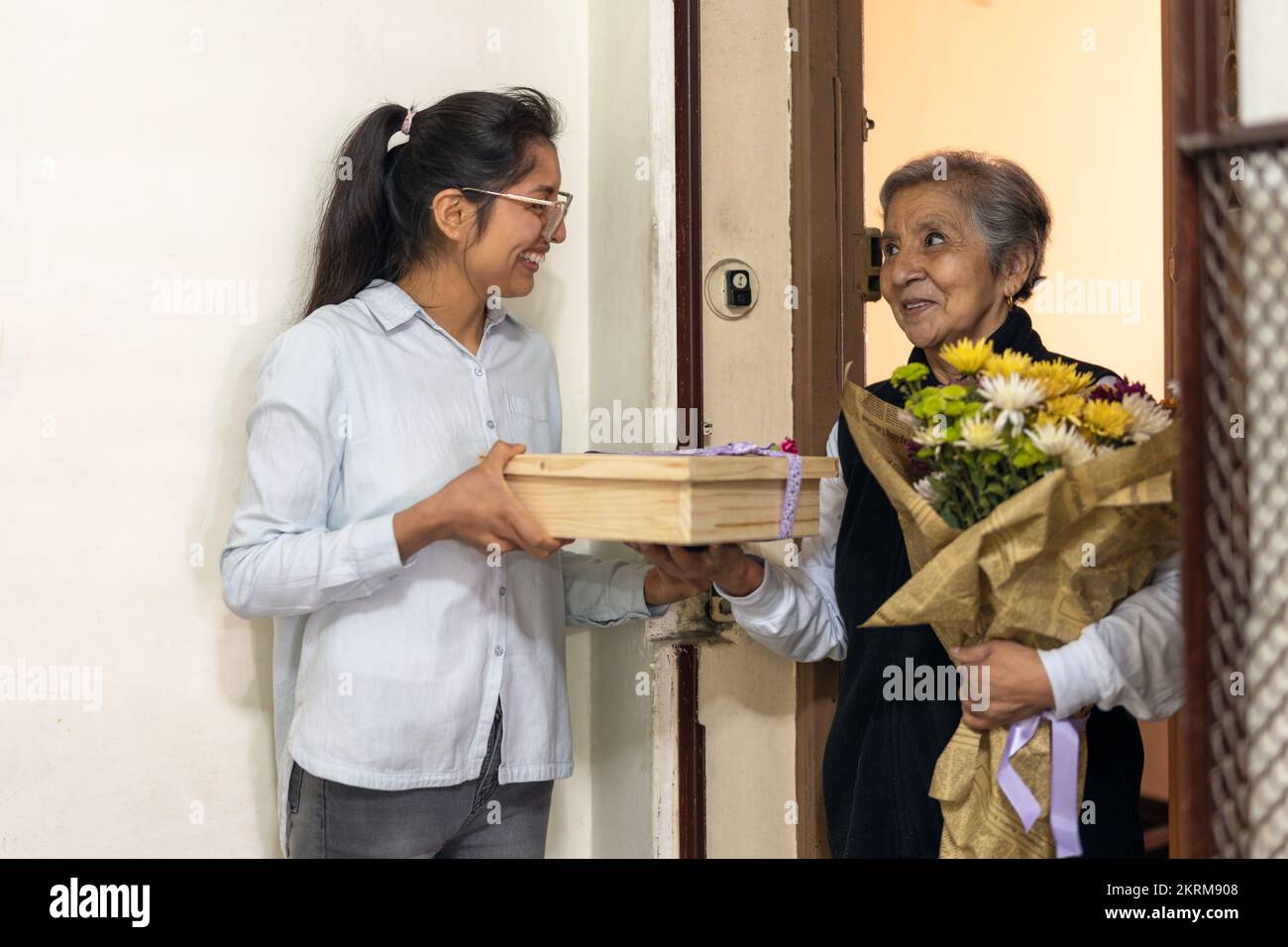 Junge Frau in lässiger Kleidung und Brille, die in der Tür steht und der Seniorin gratuliert, die einen Blumenstrauß gibt und die sich anschaut Stockfoto