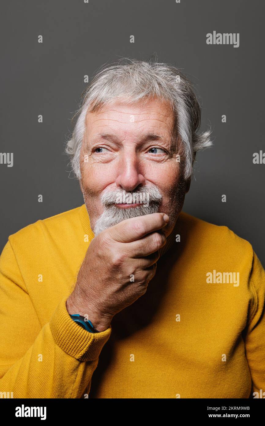 Porträt eines älteren, bärtigen, grauhaarigen Mannes in gelbem Pullover mit Armbanduhr, der mhm Gesichtsausdruck mit der Hand am Kinn macht Stockfoto