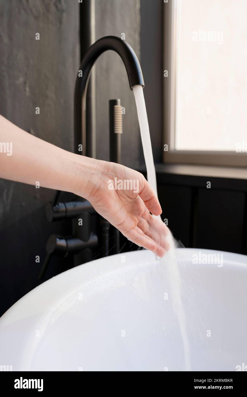 Schneiden Sie anonyme weibliche Waschhand in Badewanne, schwarzes Waschbecken, bevor Sie in einem modernen Badezimmer baden Stockfoto