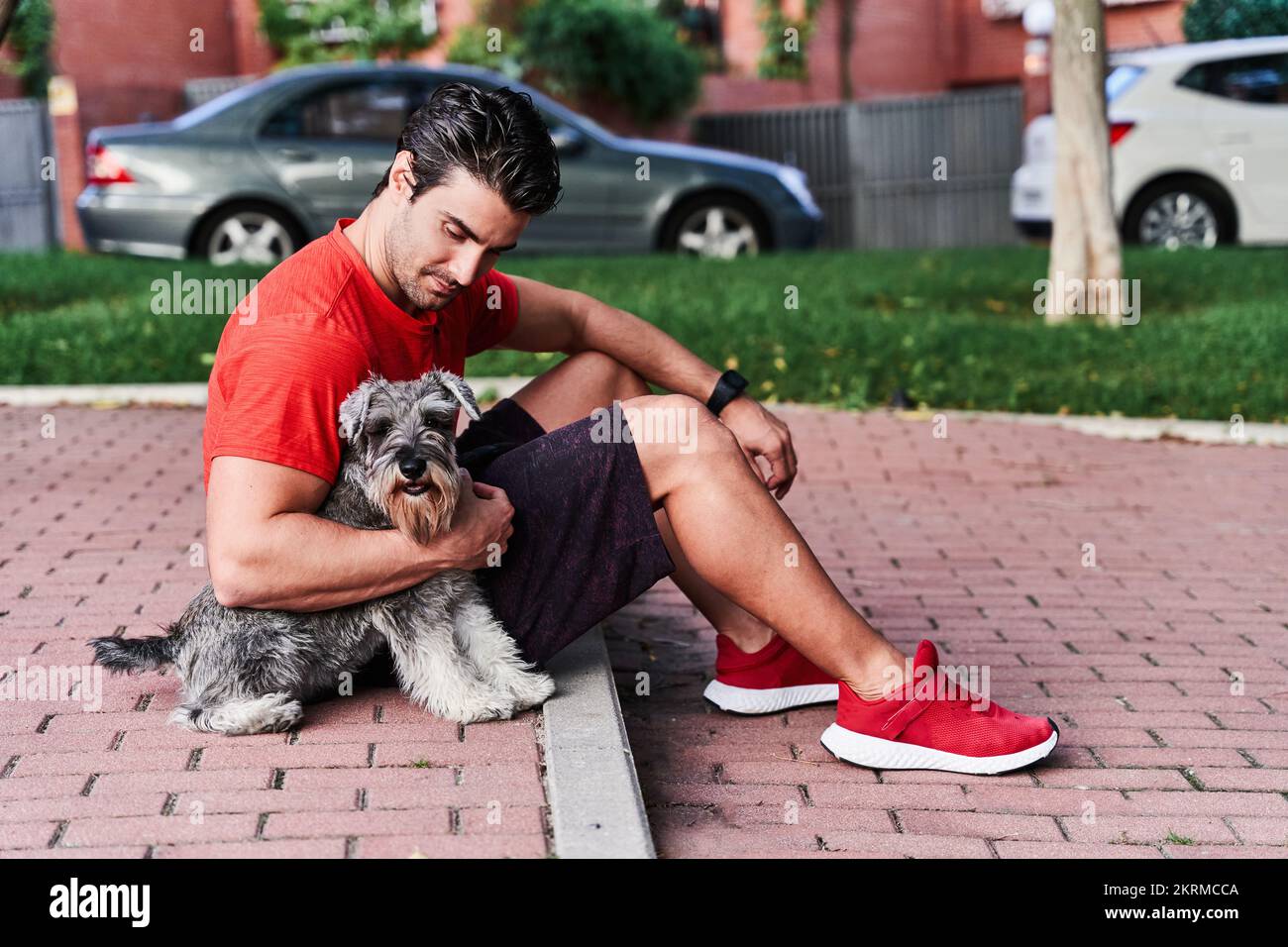 Seitenansicht des ganzen Körpers eines hispanischen Mannes in Sportbekleidung, der auf einem gepflasterten Bürgersteig sitzt und flauschigen Hund in der Nähe von Wohnhäusern umarmt Stockfoto