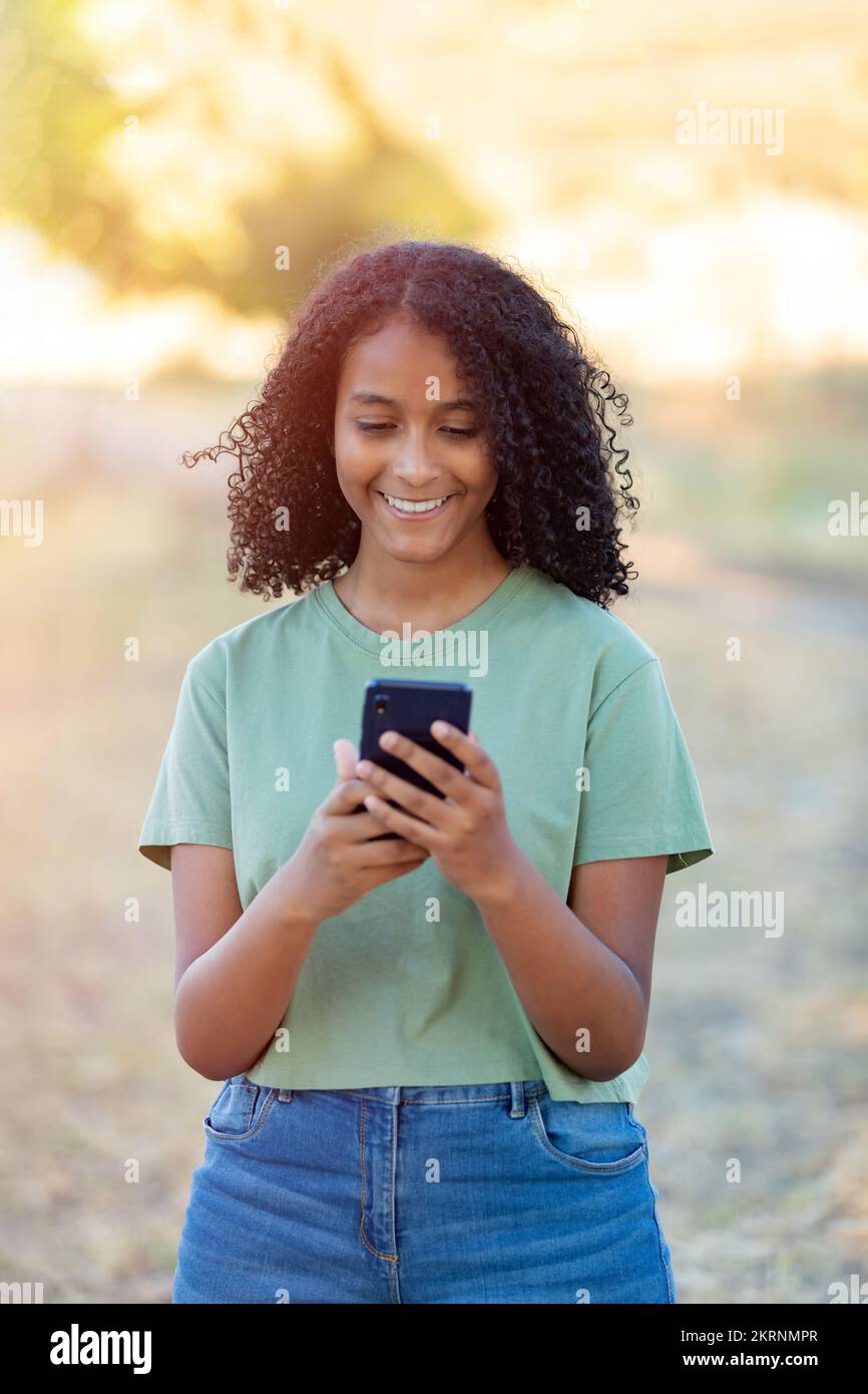 Wunderschönes lächelndes Mädchen mit lockiger Frisur, das an einem Sommertag ein Smartphone benutzt Stockfoto