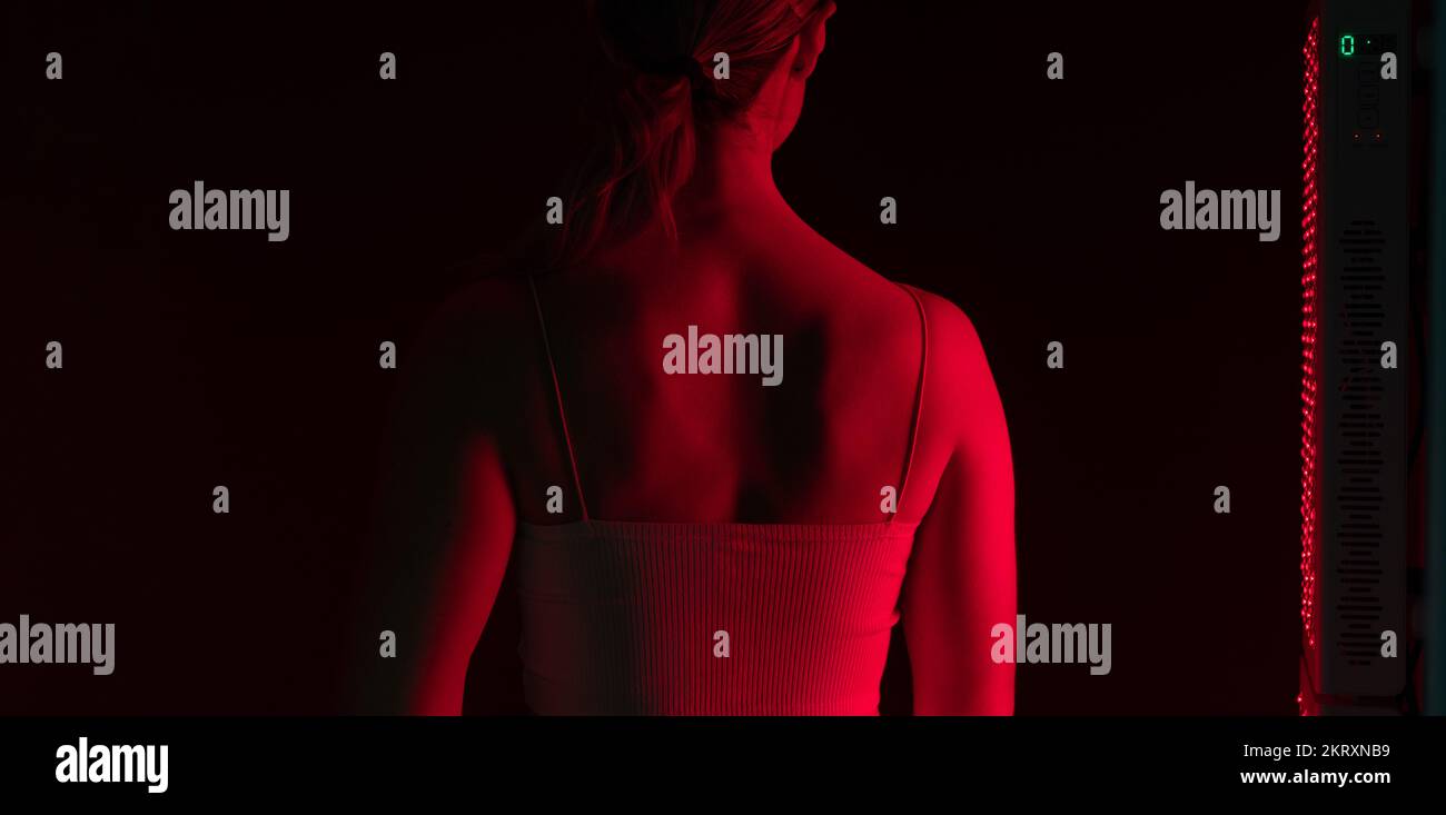 Eine Frau, die eine Rotlichttherapie auf dem Rücken bekommt. Rückblick auf eine Frau, die neben einem Rotlichtgerät in einem Schönheitssalon steht. Anti-Aging-Kosmetikbehandlung. Stockfoto