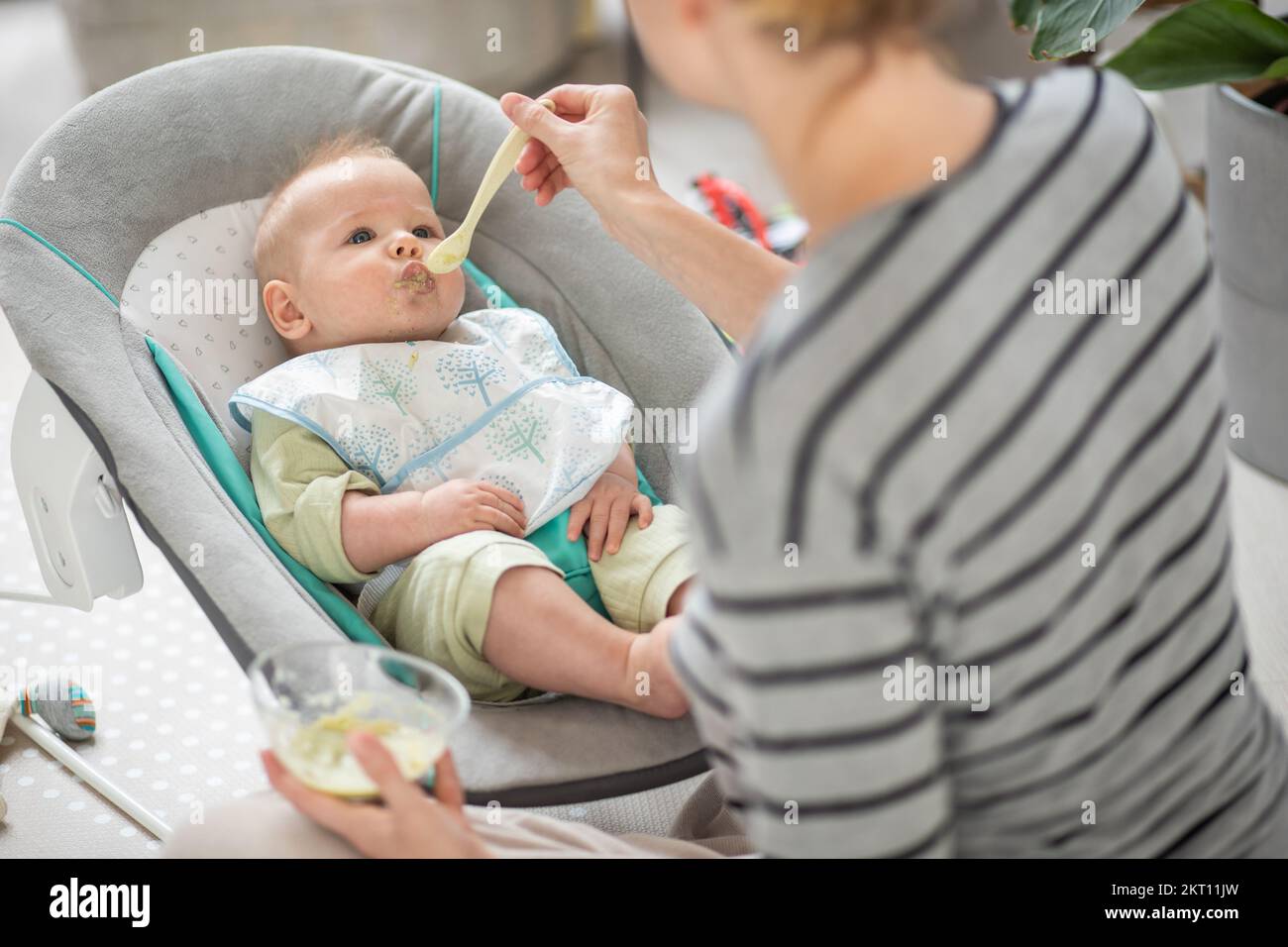 Mutter Löffel Fütterung ihres kleinen Jungen Kind in Babystuhl mit Fruchtpüree. Baby feste Nahrung Einführung Konzept Stockfoto