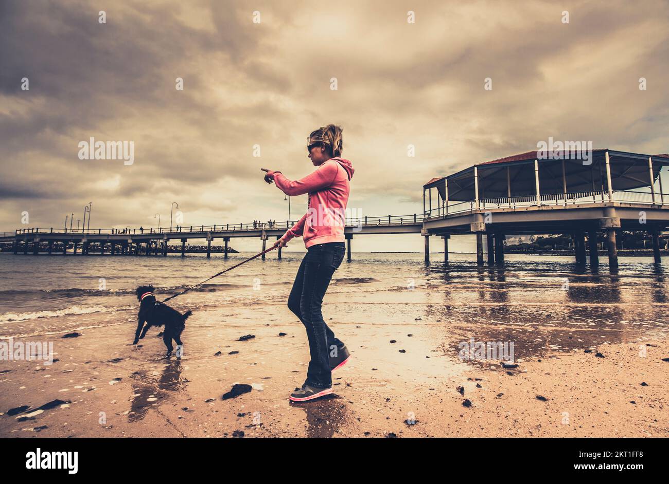 Authentisches, ehrliches instagram-Porträt einer blonden Frau Mitte der zwanziger Jahre, die zeigt, während sie den führenden Hund an der Leine führt. Redcliffe Jetty, QLD, Australien Stockfoto