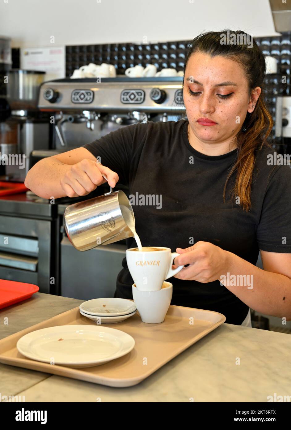 Frau, die in einem kleinen Bäckerei-Café eine Kanne heiße Milch in Kaffee gießt Stockfoto