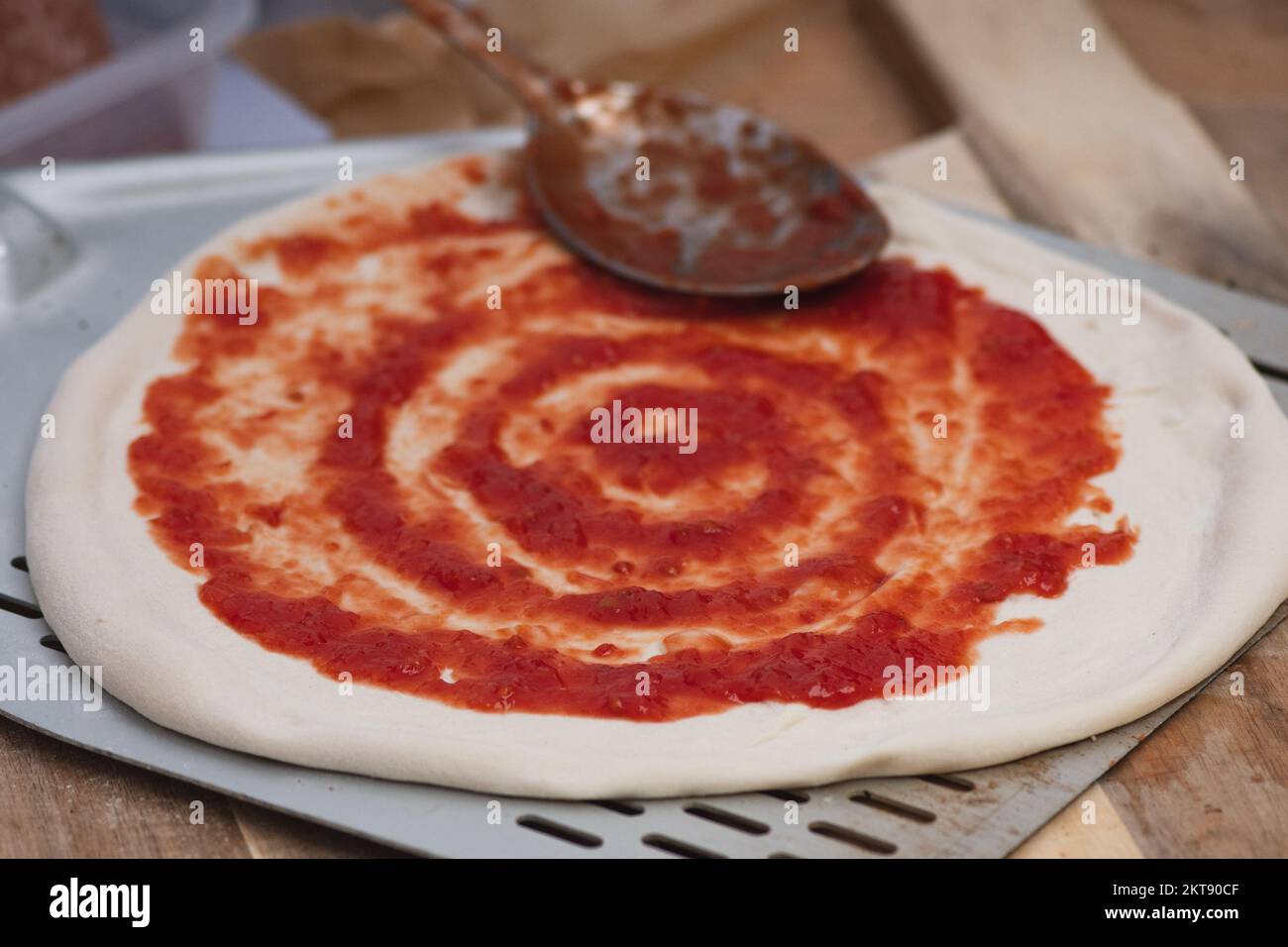 Fügen Sie Tomatensoße mit einem Löffel auf Pizzateig auf einer Schaufel auf einem Holzbrett hinzu, Nahaufnahme Stockfoto