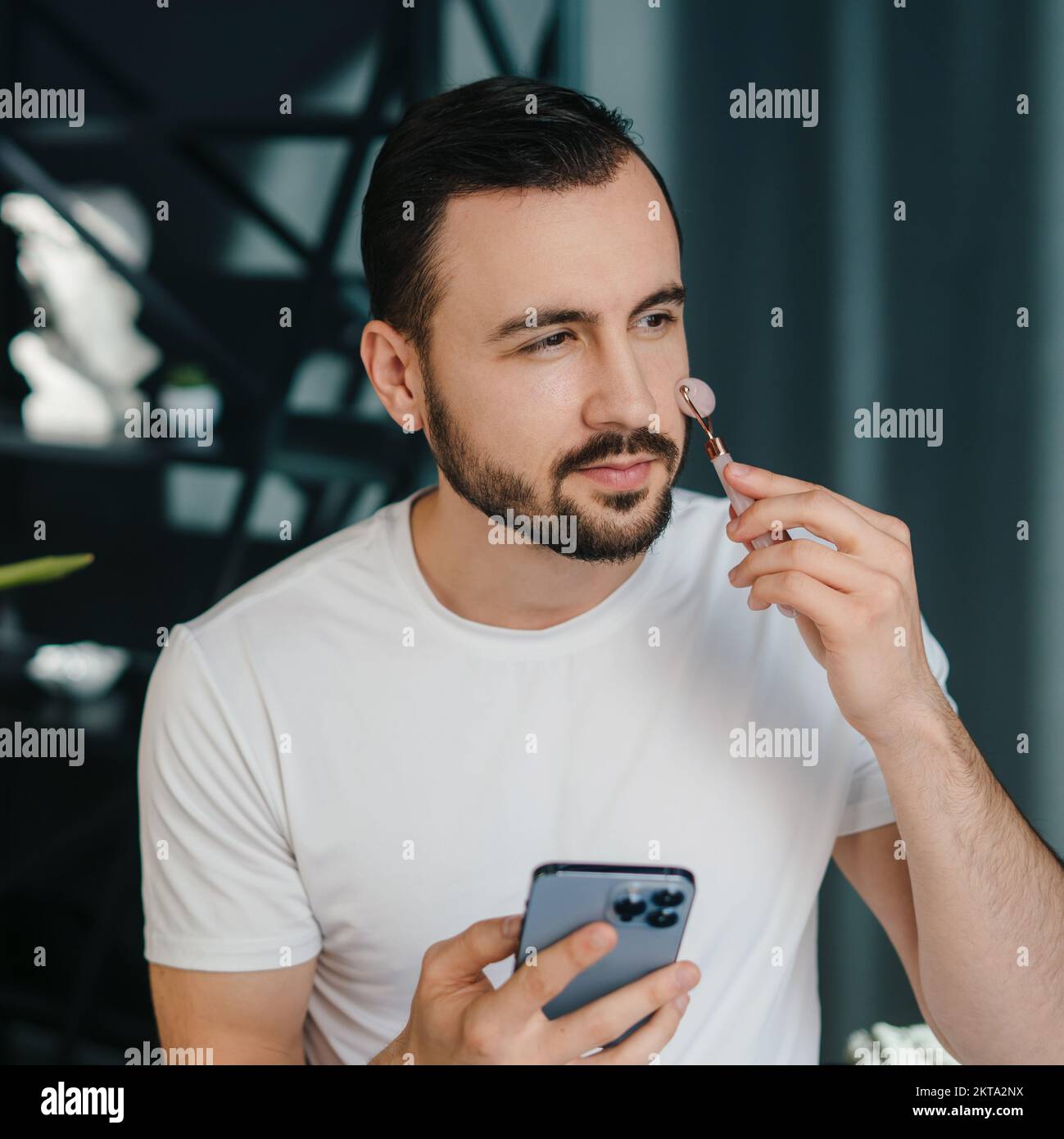 Porträt eines ruhigen Mannes, der wegschaut, während er ein Smartphone in der Hand hält und ein Massagegerät mit Jade-Rollen auf seiner Wange verwendet. Dermatologie, Kosmetologie Stockfoto