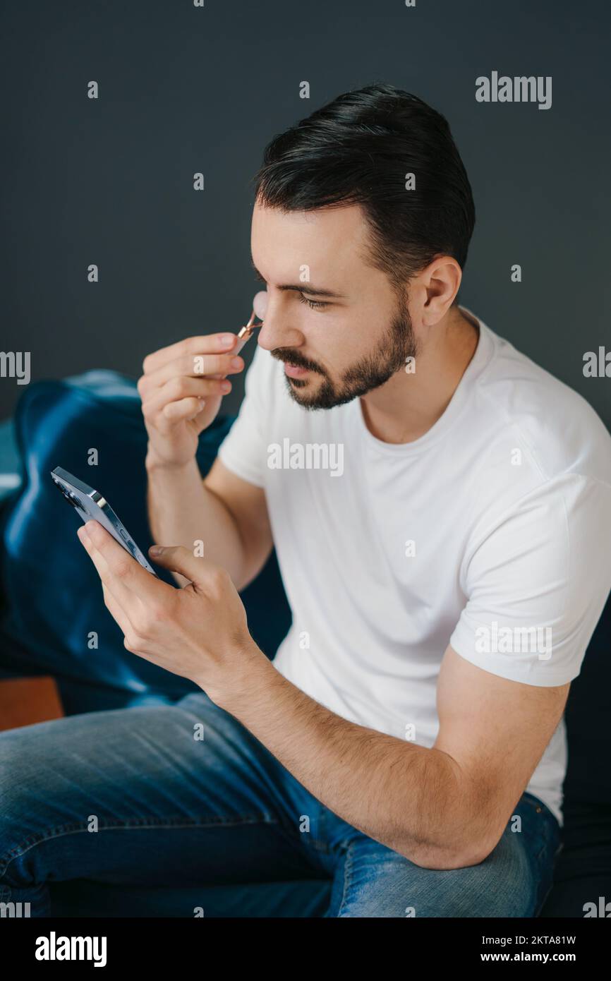Mann, der zu Hause eine Anti-Aging-Behandlung mit Jade Roller durchführt, während er auf den Smartphone-Bildschirm schaut. Dermatologie, Kosmetologie. Körperpflege. Mann Stockfoto