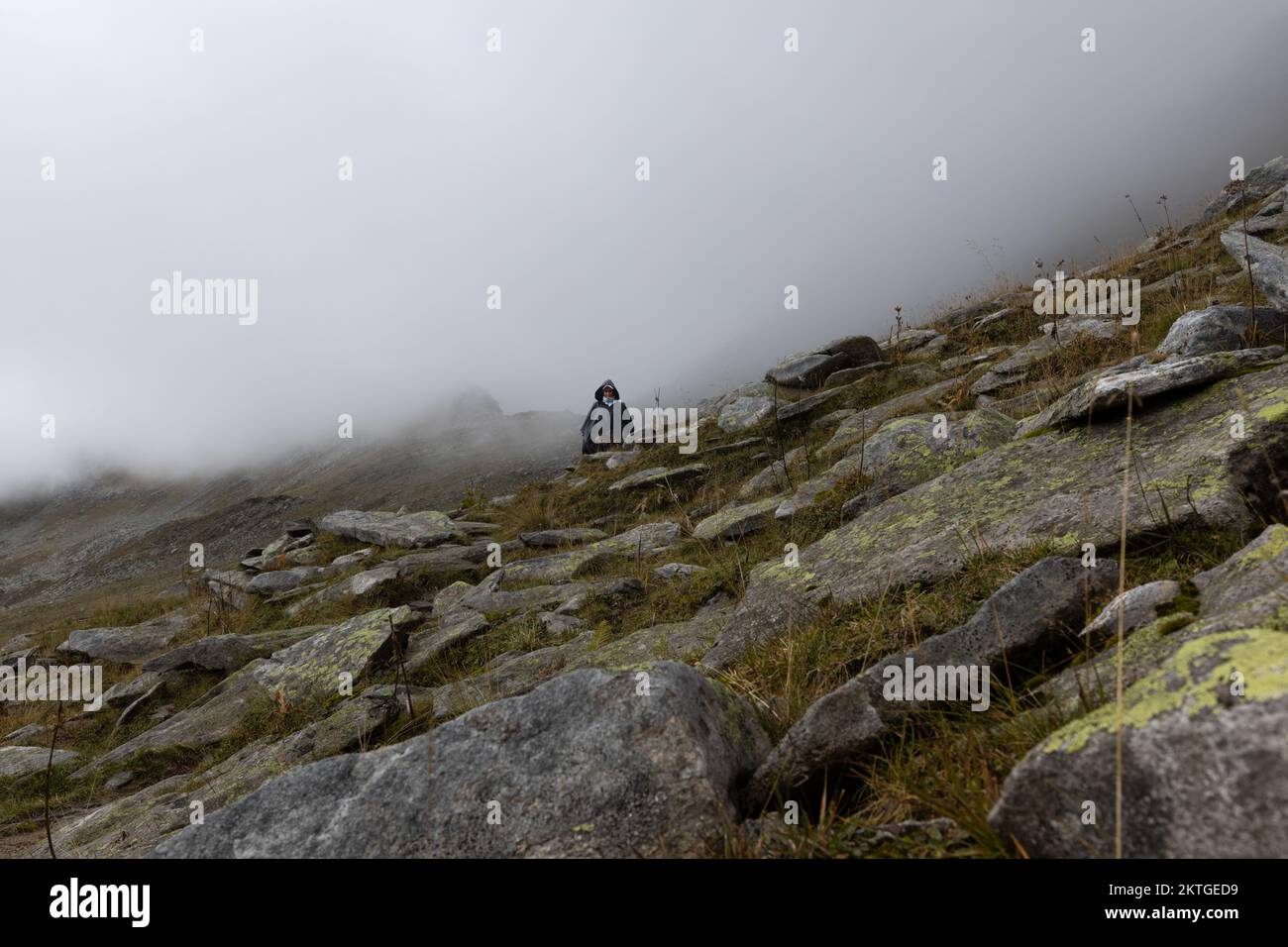 Eine Frau in einem Regenmantel geht auf einem Bergweg vor dem Hintergrund von Nebel. Österreich, Alpen. Hochwertiges Foto Stockfoto