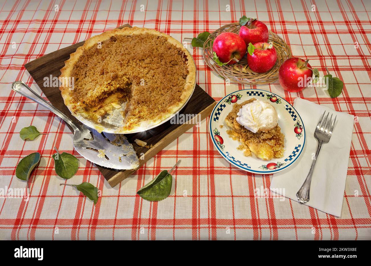 Hausgemachter Bio-Apfelkuchen als Dessert. Ein Stück Kuchen mit Eiscreme auf der Oberseite, frisch gepflückte Äpfel in einem Korb, auf roter und weißer Tischdecke. Stockfoto