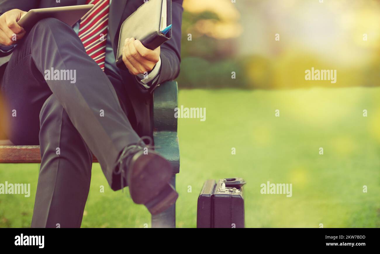 Ich mache Geschäfte im Park. Ein Geschäftsmann, der mit seinem digitalen Tablet und Organizer auf einer Parkbank sitzt. Stockfoto