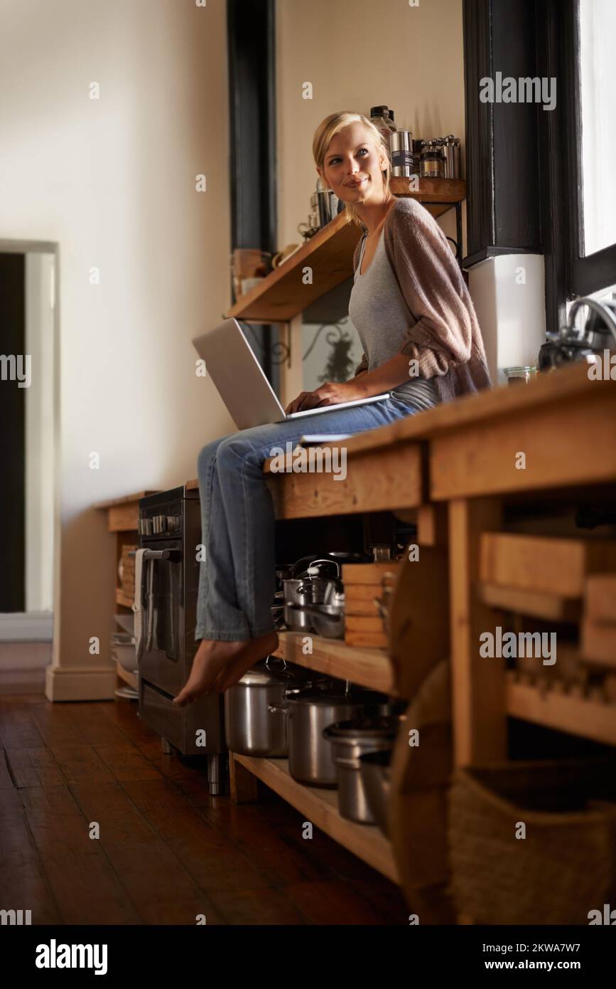 Mein Lieblingsplatz zum Arbeiten. Porträt einer attraktiven jungen Frau, die ihren Laptop benutzt, während sie sich zu Hause auf ihrer Küchentheke entspannt. Stockfoto