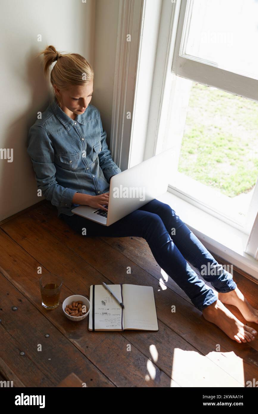 Weg vom Trubel des Zuhauses. Eine junge Frau, die am Fenster auf dem Boden sitzt und etwas über ihren Laptop recherchiert. Stockfoto