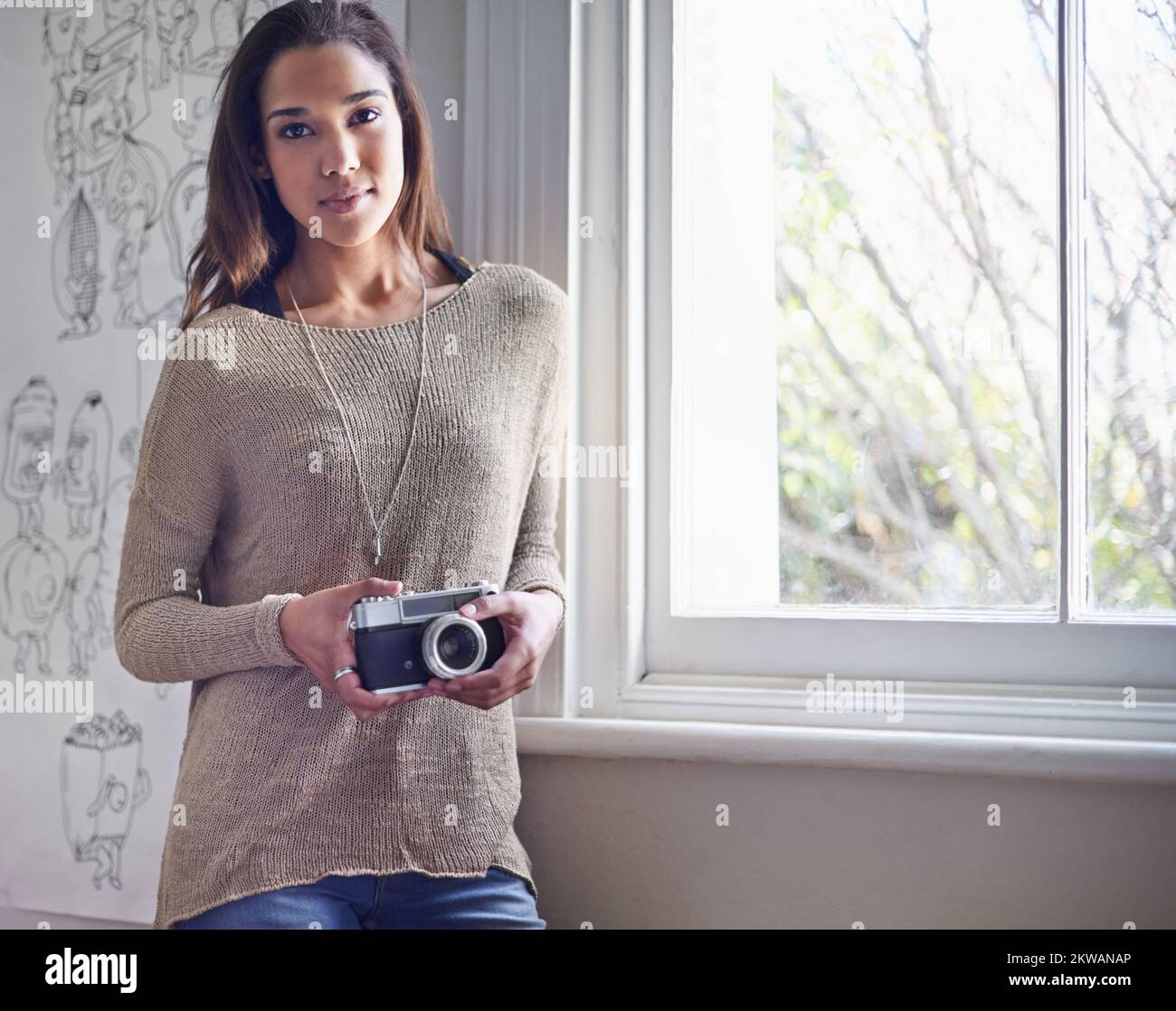 Sie liebt Fotografie. Eine junge Fotografin, die neben einem Fenster steht, während sie ihre Kamera hält. Stockfoto