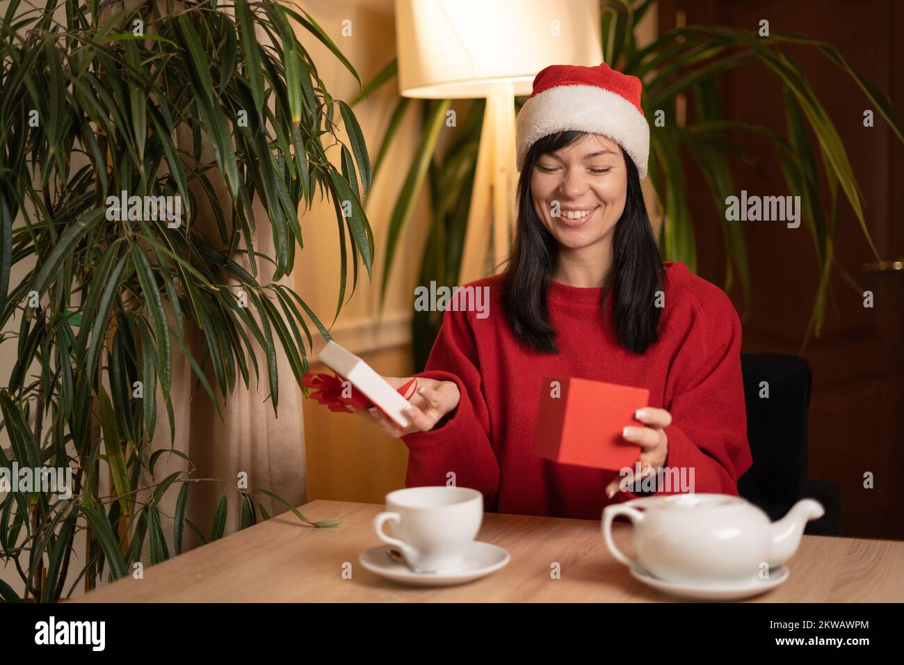 Junge, lachende brünette Frau mit weihnachtsmannmütze öffnet eine Schachtel mit einem Geschenk, lächelnd zufrieden mit einem guten Weihnachtsgeschenk. Speicherplatz kopieren Stockfoto