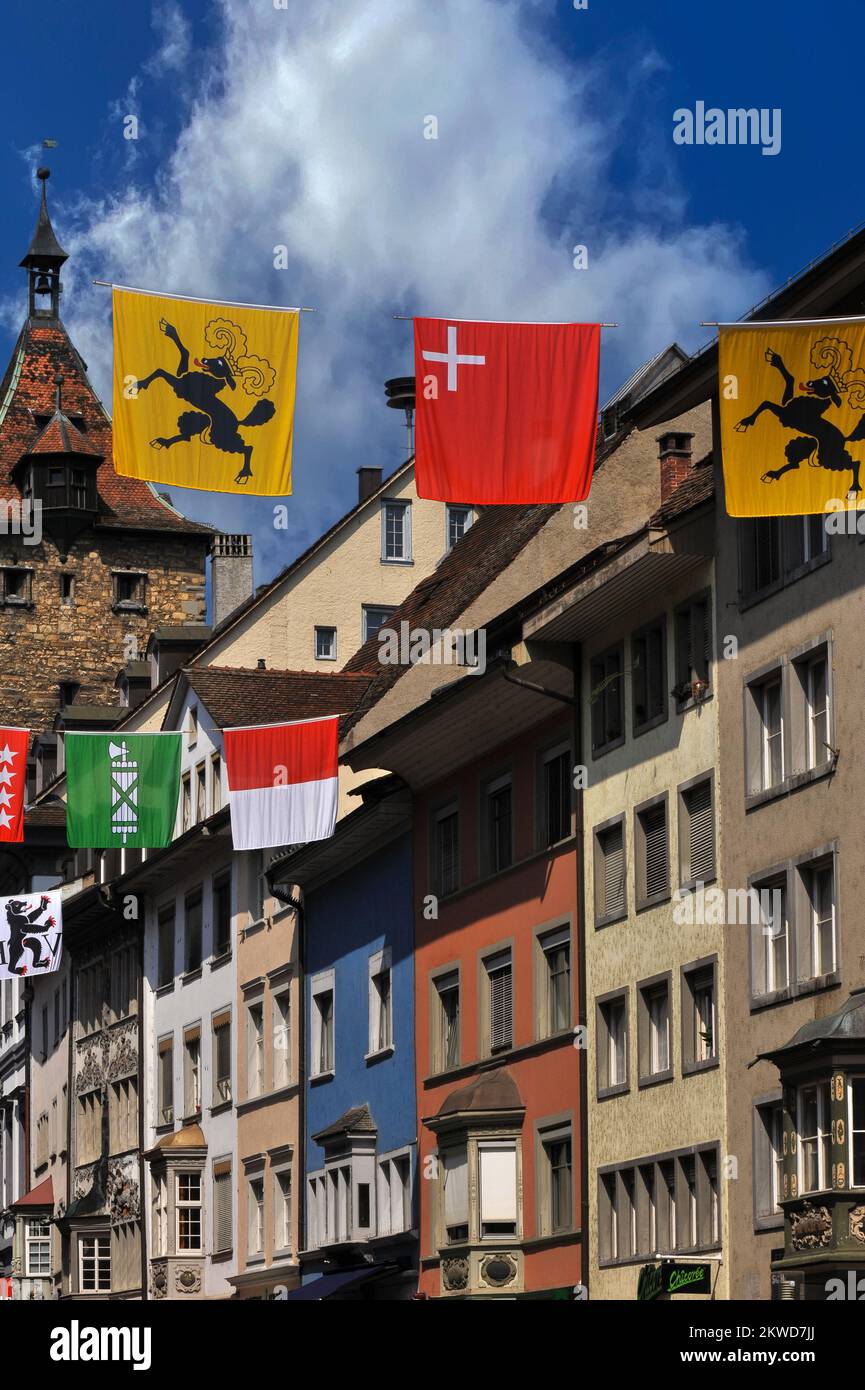 Lebhafte Banner oder Flaggen der 26 Schweizer Kantone hängen zwischen Renaissance- und barocken ehemaligen Handelshäusern mit Orielfenstern in der Vordergasse in der Altstadt von Schaffhausen, Nordschweiz. Dahinter befindet sich der mittelalterliche Obertor Turm, der ehemalige westliche Eingang zur Stadt und der älteste noch erhaltene Teil der Befestigungsanlagen. Stockfoto